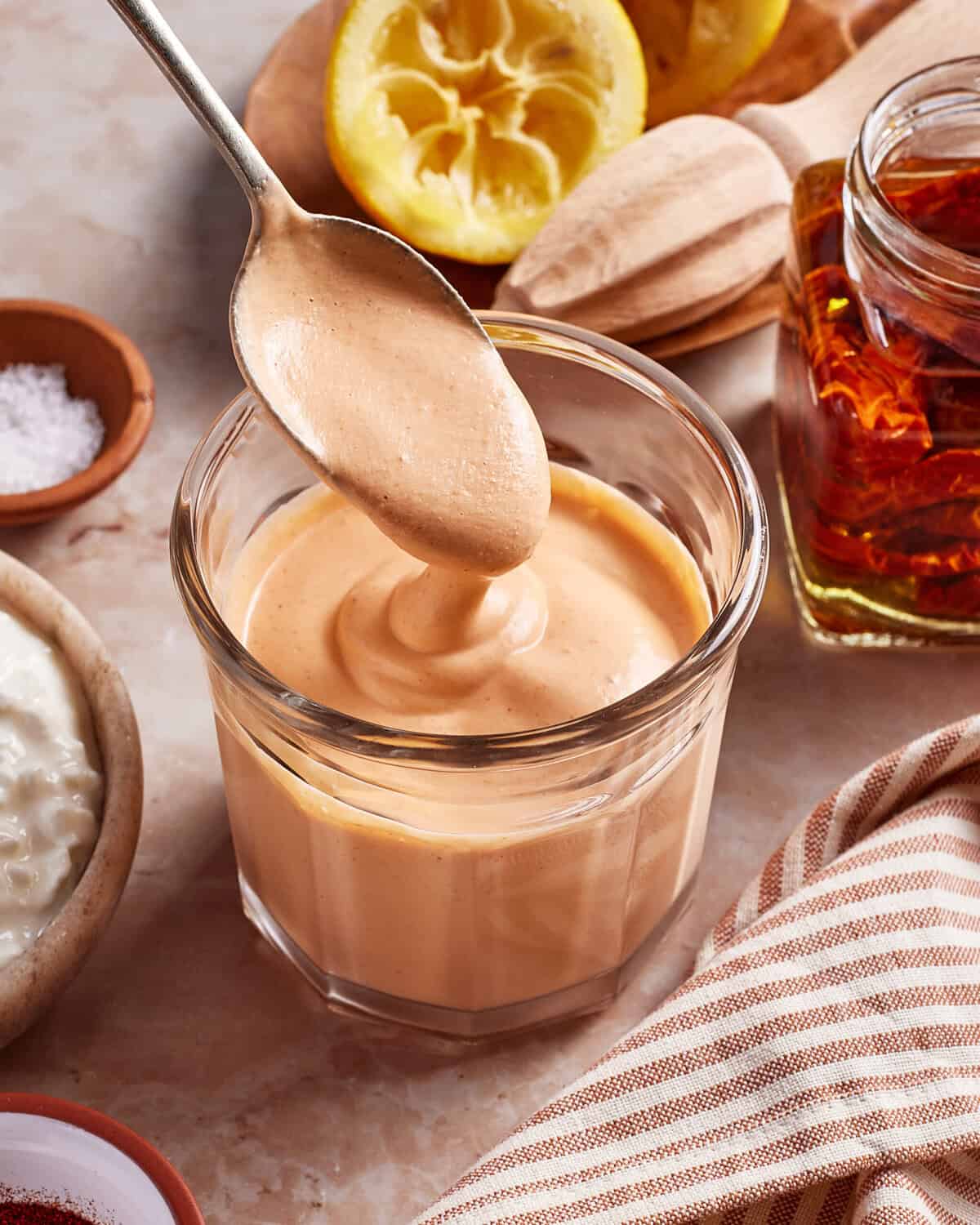 A spoon drizzles creamy, light brown sauce into a glass jar. In the background are a halved lemon, wooden juicer, a jar of oil with chili, salt, and a striped cloth.