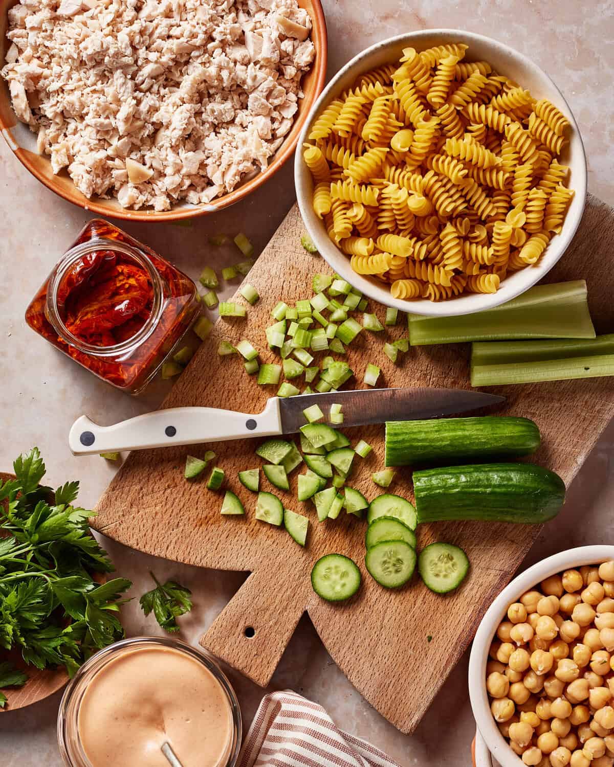 A wooden cutting board with chopped cucumber and celery, a knife, and surrounded by bowls of rotini pasta, cooked chicken, chickpeas, parsley, sundried tomatoes, and a jar of creamy dressing.