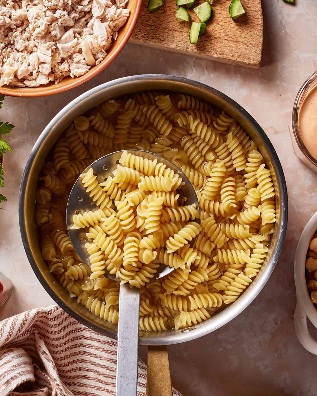 A pot of cooked rotini pasta with a large serving spoon, surrounded by chopped cucumbers, shredded chicken, and a bowl of dressing on a marble countertop.