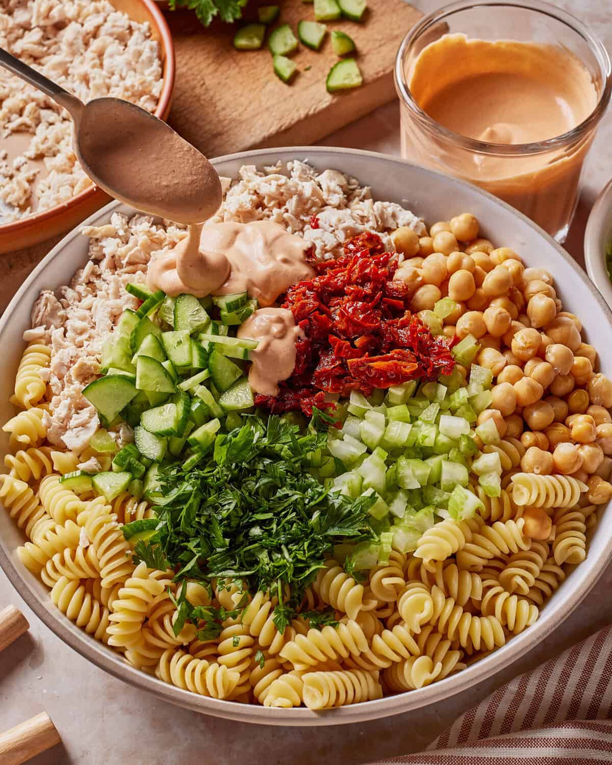 A bowl filled with rotini pasta, chickpeas, cucumber, sun-dried tomatoes, celery, fresh herbs, and shredded chicken, with a spoon drizzling creamy dressing on top. A glass of dressing is nearby. High Protein Tuscan Chicken Pasta Salad