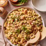 A large bowl of High Protein Tuscan Chicken Pasta Salad with vegetables and fresh parsley, served with wooden salad utensils. Surrounding the bowl are a napkin, a small dish of salt, a bunch of parsley, pepper grinder, and a jar of dressing.