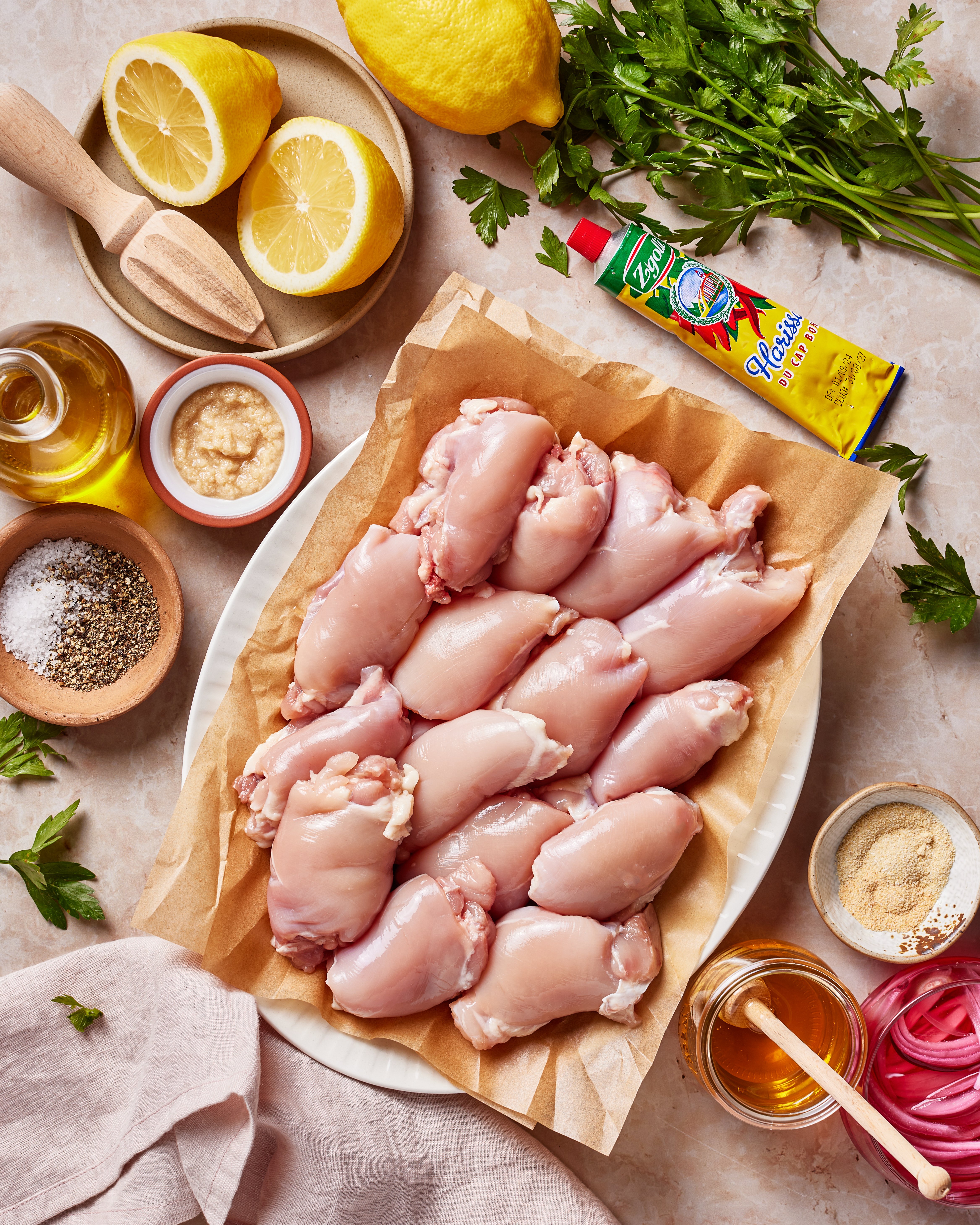 Raw chicken thighs on parchment paper surrounded by lemon halves, parsley, olive oil, mustard, tomato paste, salt, pepper, sliced onions, and seasonings on a light countertop.