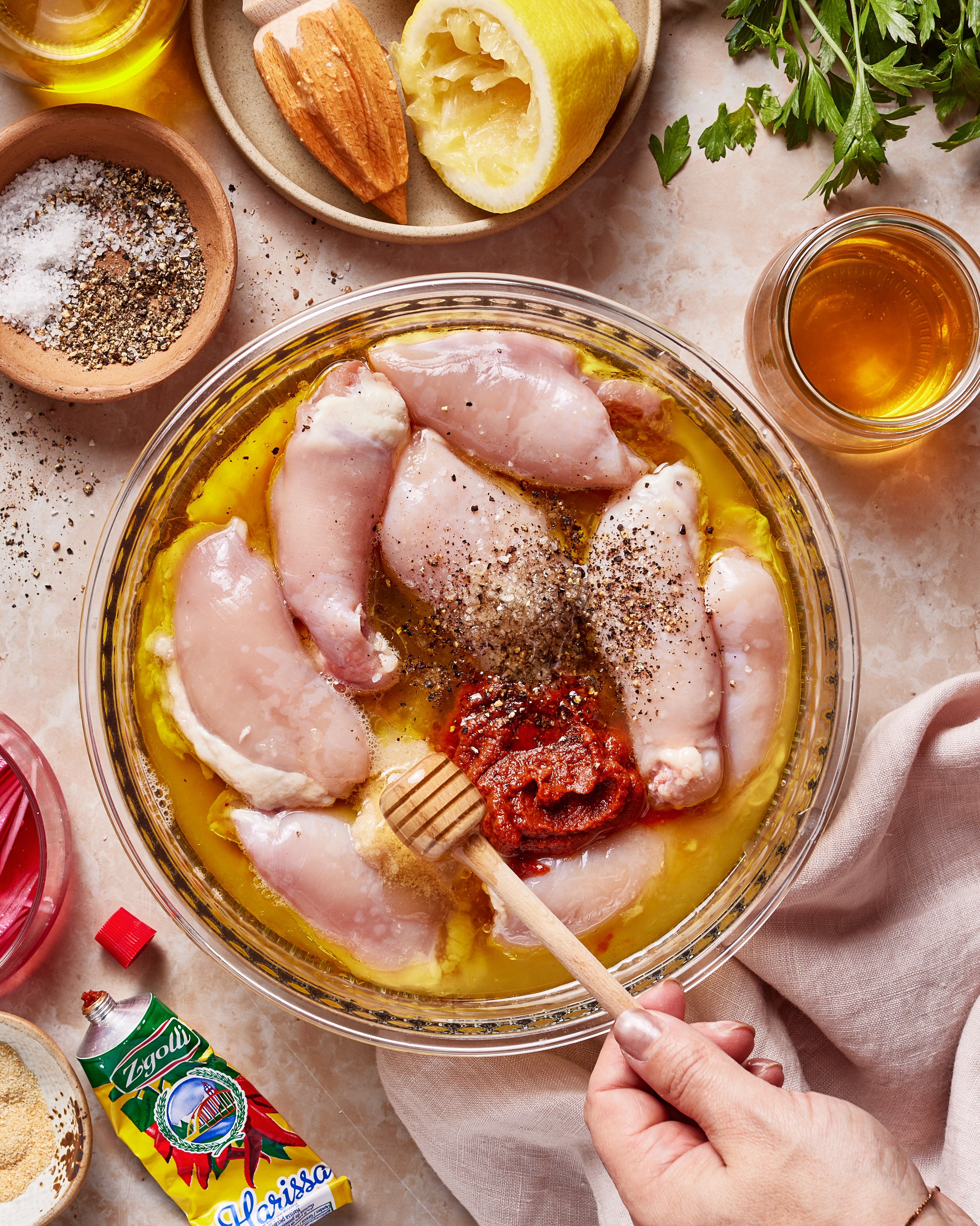 A hand uses a honey dipper to mix raw chicken in a bowl with oil, spices, and harissa paste. Surrounding items include salt, pepper, lemon, parsley, bread, olive oil, and a packet of harissa paste.