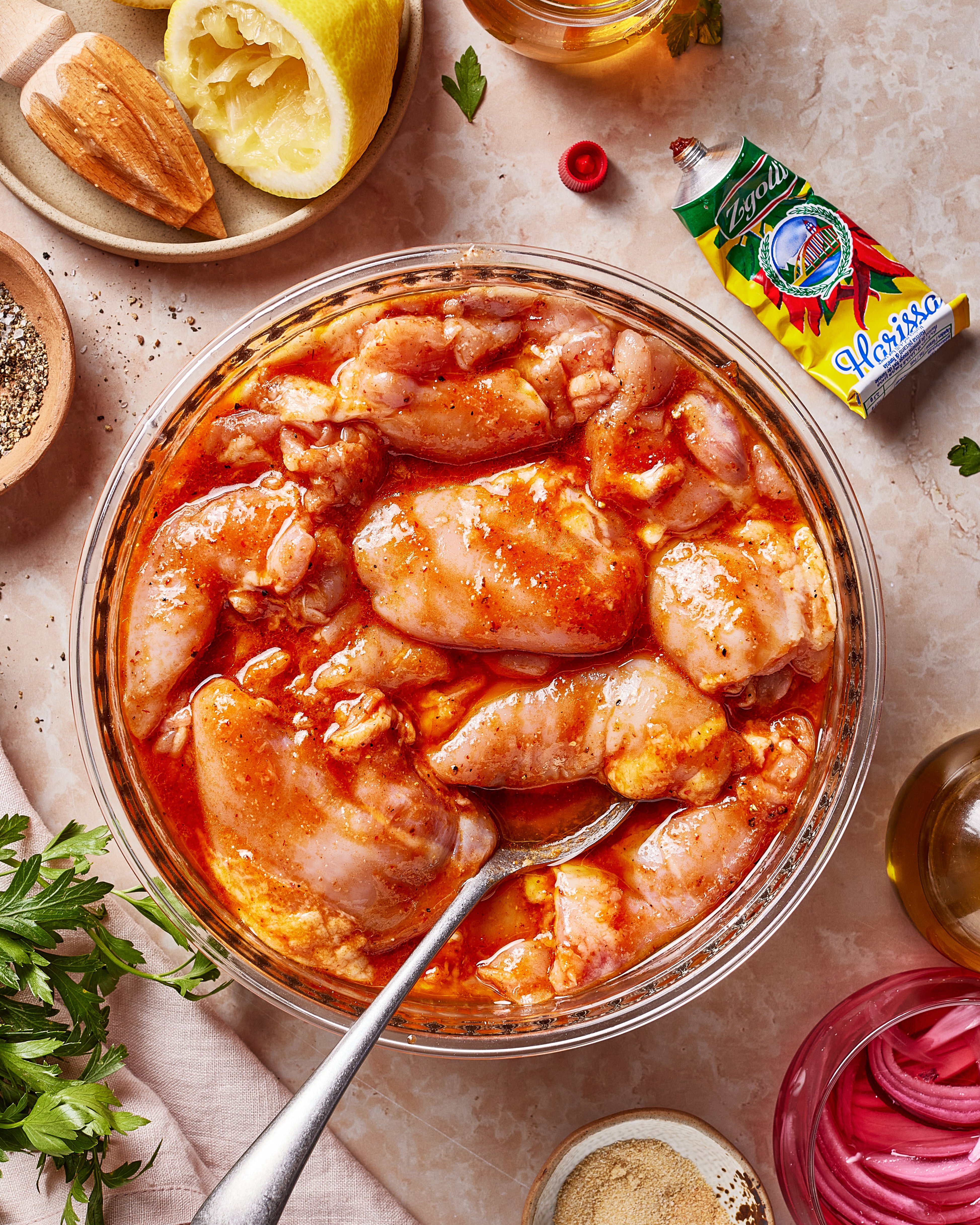 Raw chicken pieces marinating in a bowl with red sauce, surrounded by spices, parsley, lemon, oil, and a packet of harissa on a marble surface; a spoon rests in the bowl.