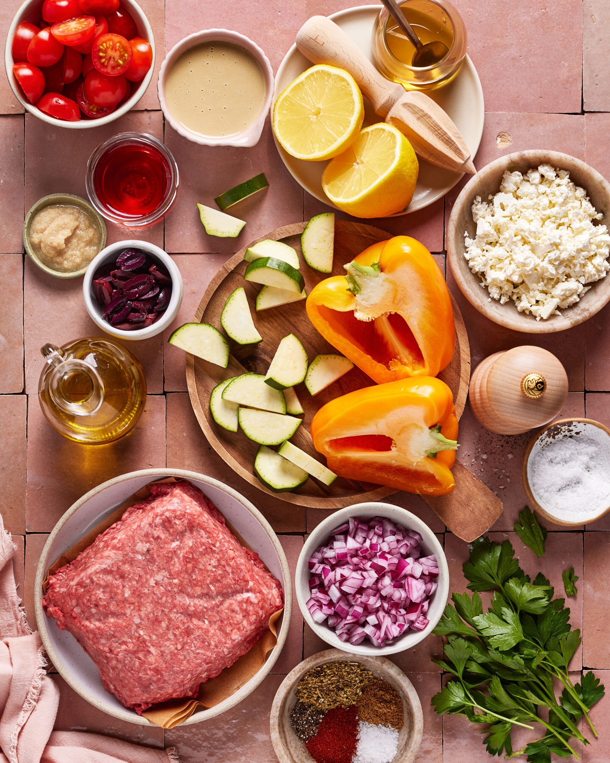 Top-down view of ingredients on a pink tile surface, including ground meat, yellow bell peppers, zucchini, red onion, grape tomatoes, feta cheese, parsley, olive oil, seasonings, lemon, and small bowls of condiments.