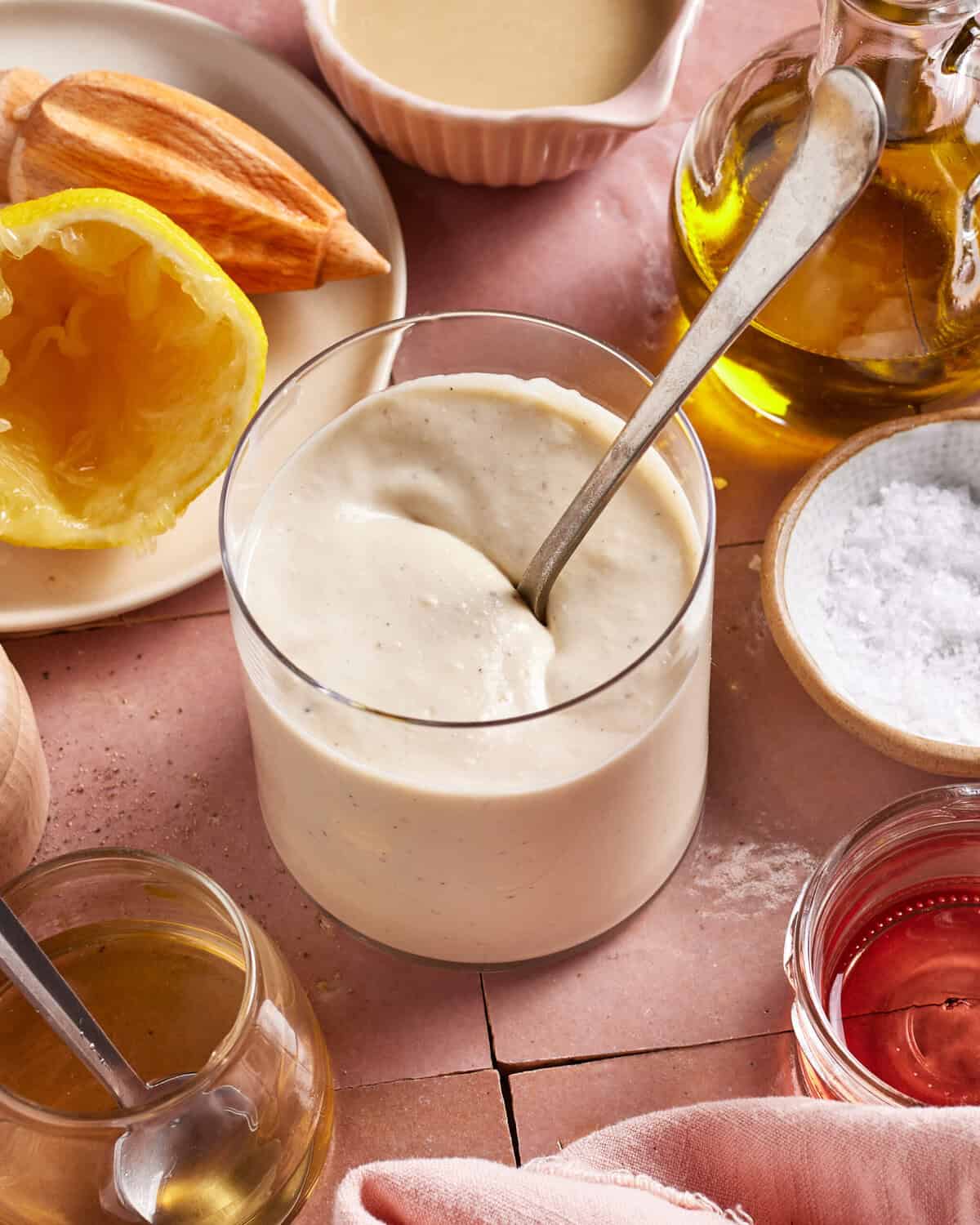A glass filled with creamy homemade mayonnaise sits on a pink surface, surrounded by a halved lemon, olive oil, salt, vinegar, mustard, and a wooden citrus juicer. A spoon rests in the mayonnaise.