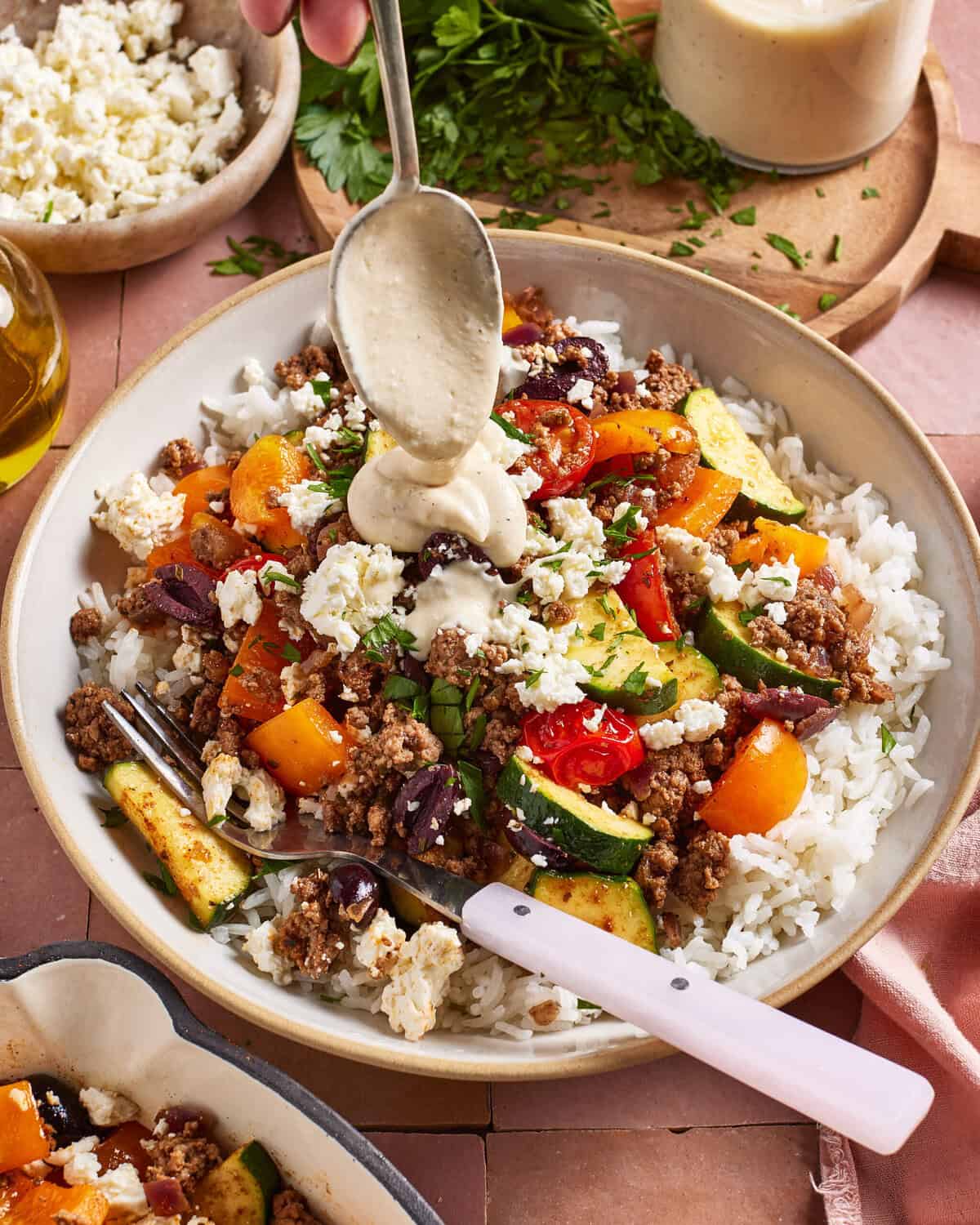 A bowl of rice topped with ground meat, zucchini, tomatoes, bell peppers, feta cheese, and olives, with a spoon drizzling sauce on top. Fresh herbs, cheese, and sauce are visible in the background.