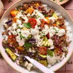 A bowl of rice topped with ground meat, cherry tomatoes, zucchini, bell peppers, kalamata olives, feta cheese, fresh parsley, and a drizzle of creamy sauce, with a fork on the side.