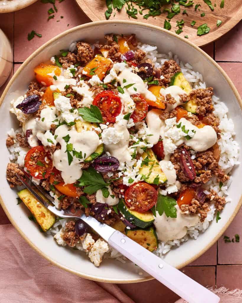 A bowl of rice topped with ground meat, cherry tomatoes, zucchini, bell peppers, kalamata olives, feta cheese, fresh parsley, and a drizzle of creamy sauce, with a fork on the side.