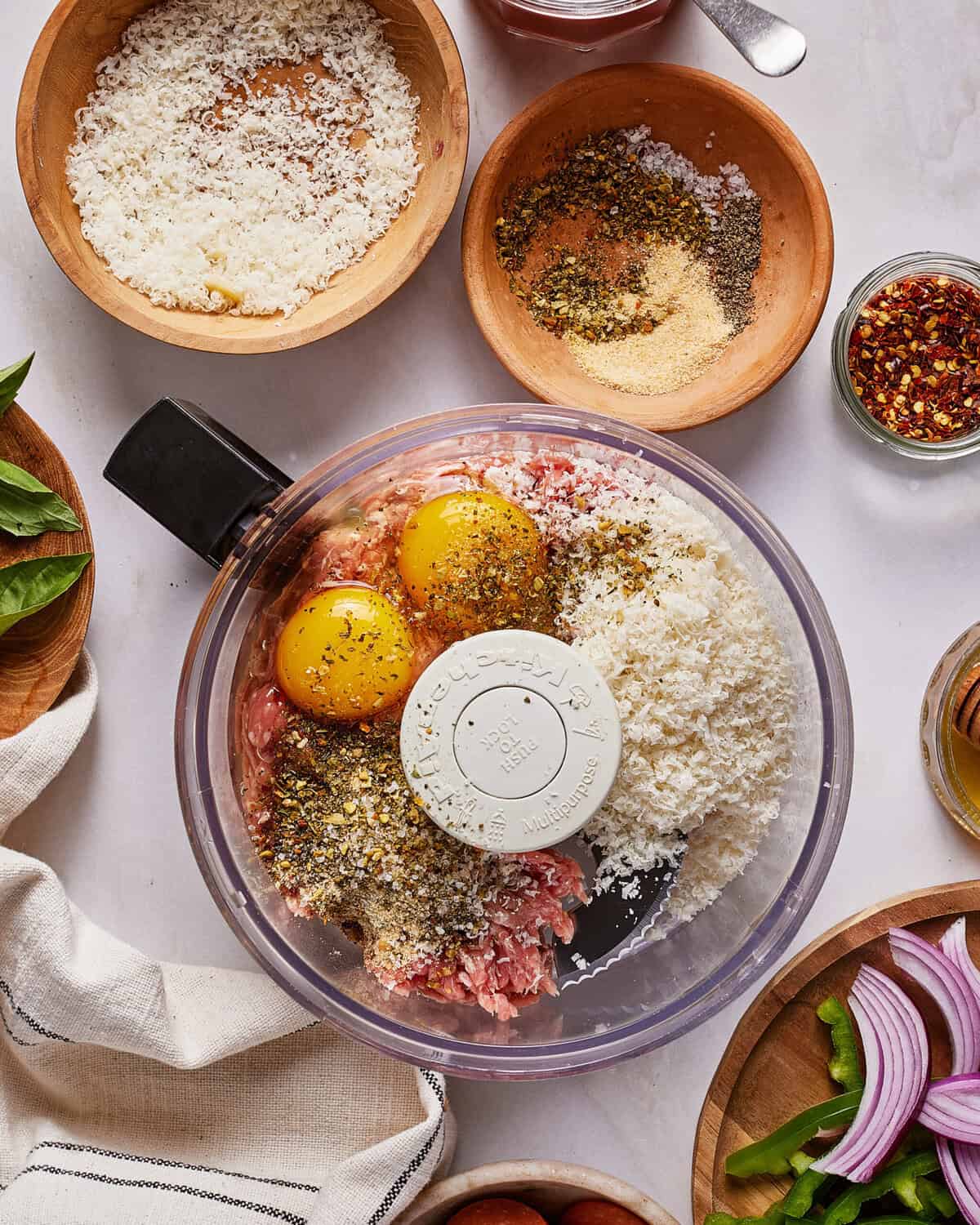 A food processor bowl filled with ground meat, two eggs, breadcrumbs, grated cheese, and herbs sits on a table surrounded by bowls of spices, cheese, and sliced onions.