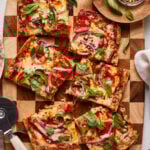 Six rectangular slices of pizza with melted cheese, pepperoni, red onion, and fresh basil on a wooden board. A pizza cutter, small bowl of red pepper flakes, and some extra basil are nearby.