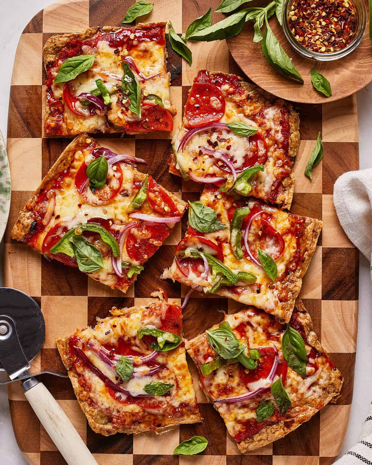 Six rectangular slices of pizza with melted cheese, pepperoni, red onion, and fresh basil on a wooden board. A pizza cutter, small bowl of red pepper flakes, and some extra basil are nearby.