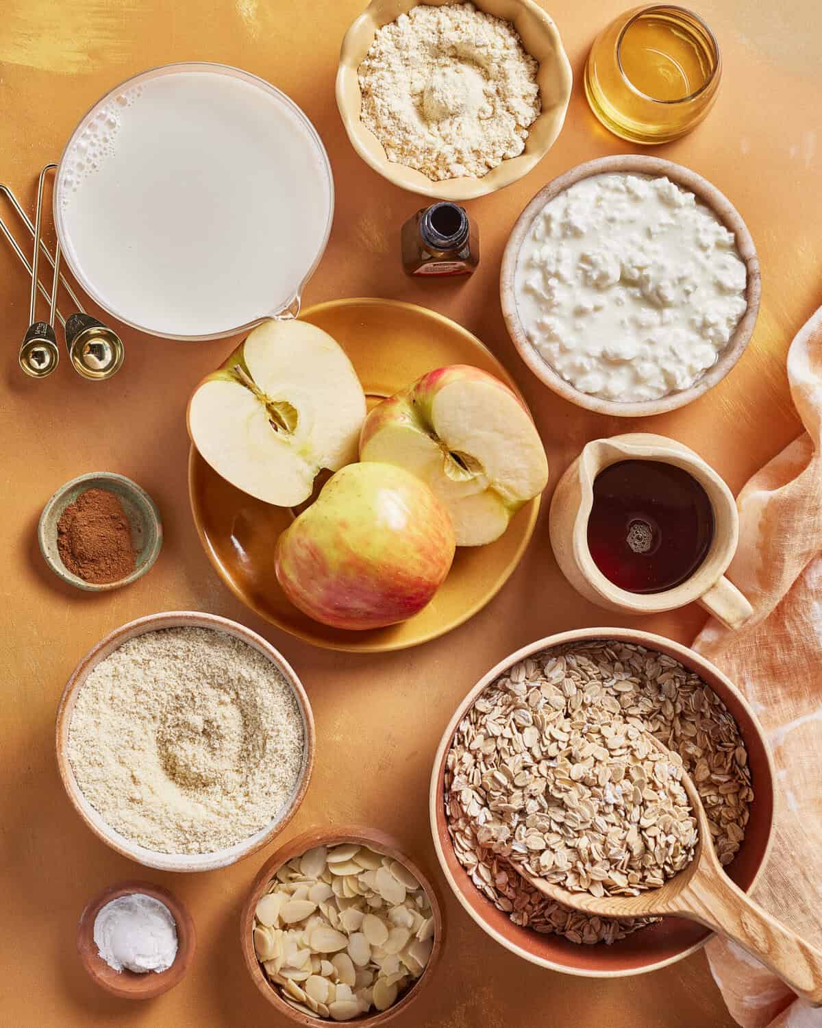 A variety of baking ingredients on a light orange surface, including oats, sliced apples, cottage cheese, almond flour, sliced almonds, milk, honey, ground cinnamon, maple syrup, vanilla, and small measuring spoons.