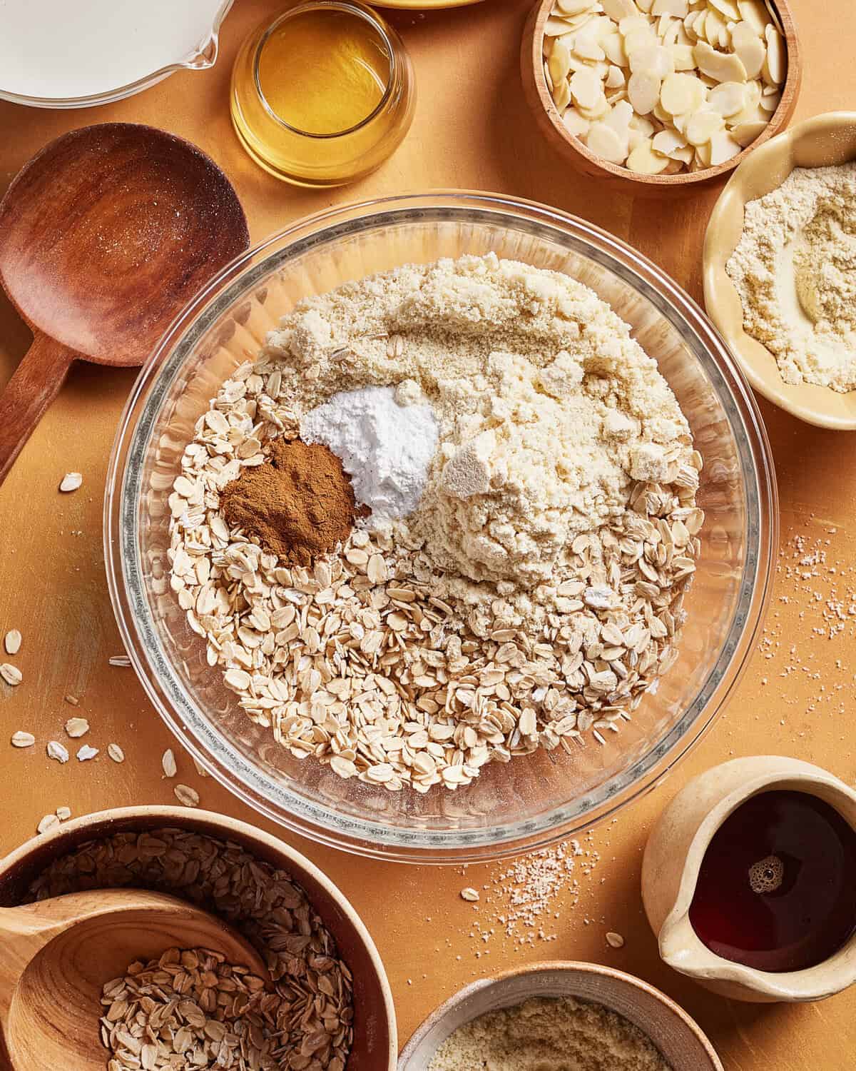 A glass bowl contains rolled oats, almond flour, baking powder, and cinnamon. Surrounding it are bowls with sliced almonds, flour, oats, maple syrup, milk, and a wooden spoon on an orange surface.