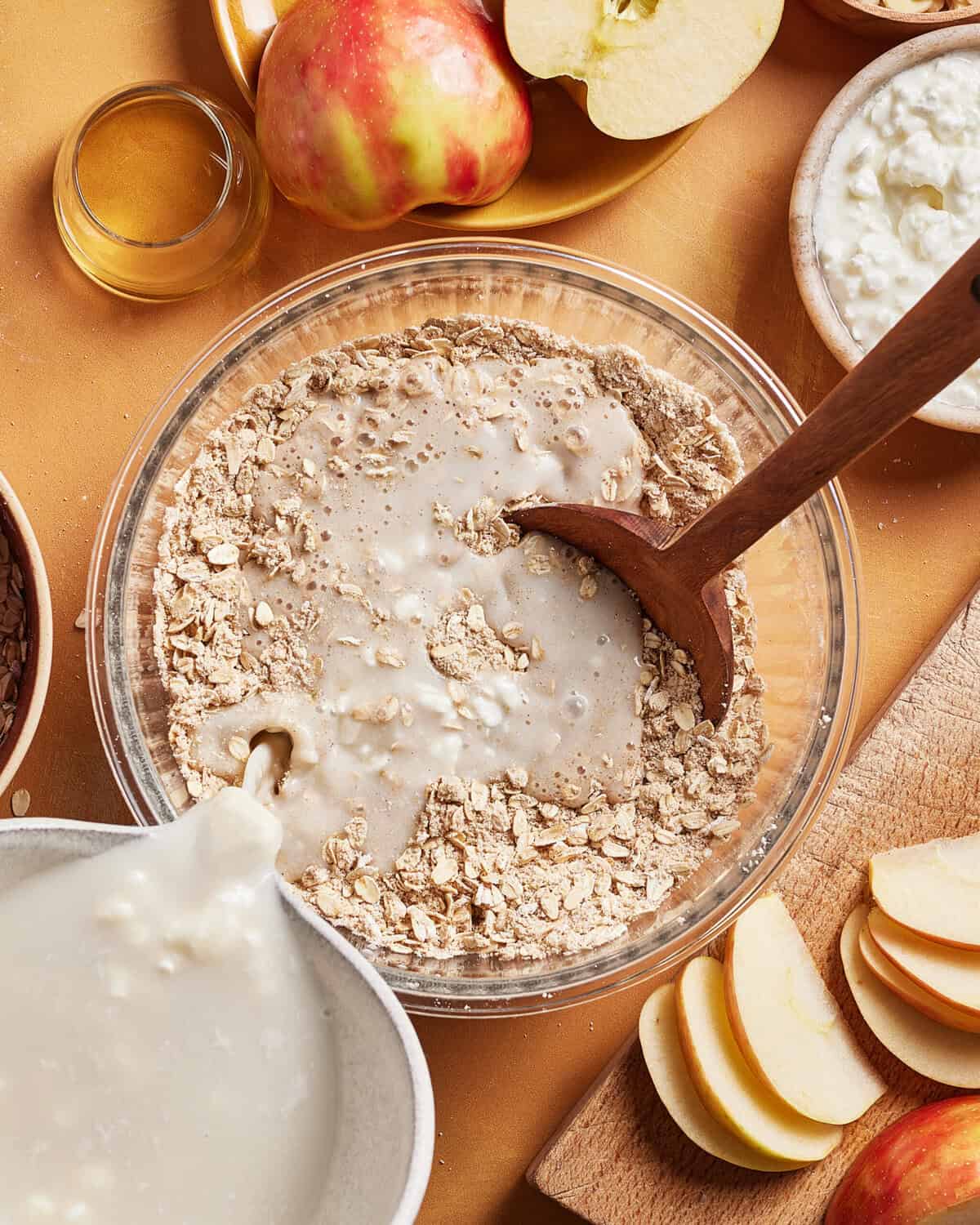 A bowl of dry oats being mixed with milk using a wooden spoon, surrounded by sliced apples, cottage cheese, a small glass of honey, and other ingredients on a table.