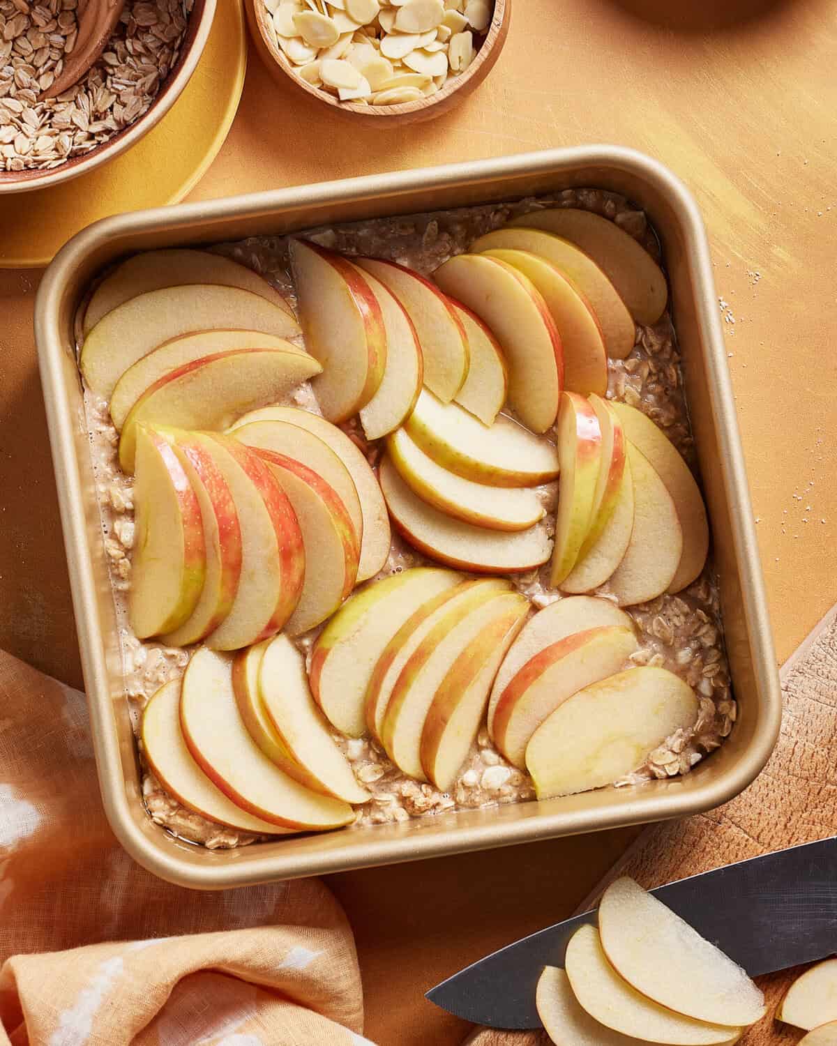 A square baking dish filled with oats and topped with neatly arranged slices of red and yellow apples. A small bowl of sliced almonds, a bowl of oats, and a knife are nearby on a golden-colored surface.