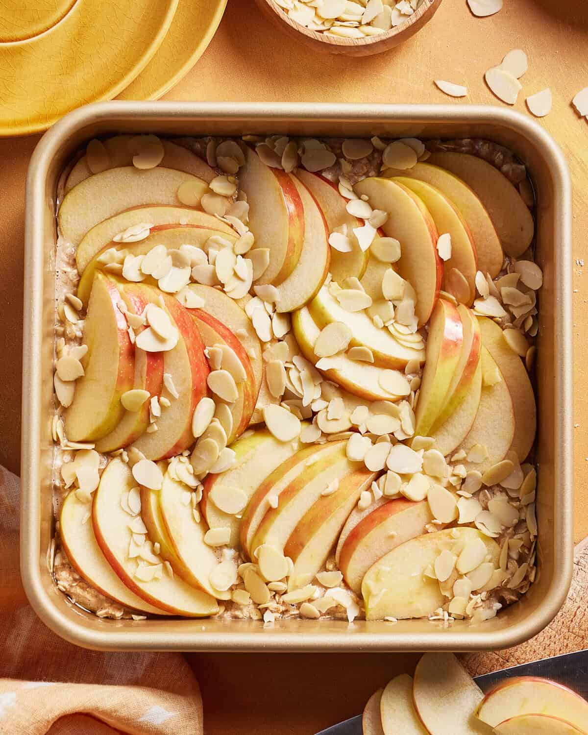 A square baking pan filled with neatly arranged apple slices, topped with slivered almonds, sits on a wooden surface with plates and a bowl of extra almonds nearby.