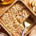 A square baking dish of sliced apple and almond dessert, with three pieces cut and a serving spatula inside. Surrounding the dish are plates, apple slices, and a bowl of oats on a light brown surface.