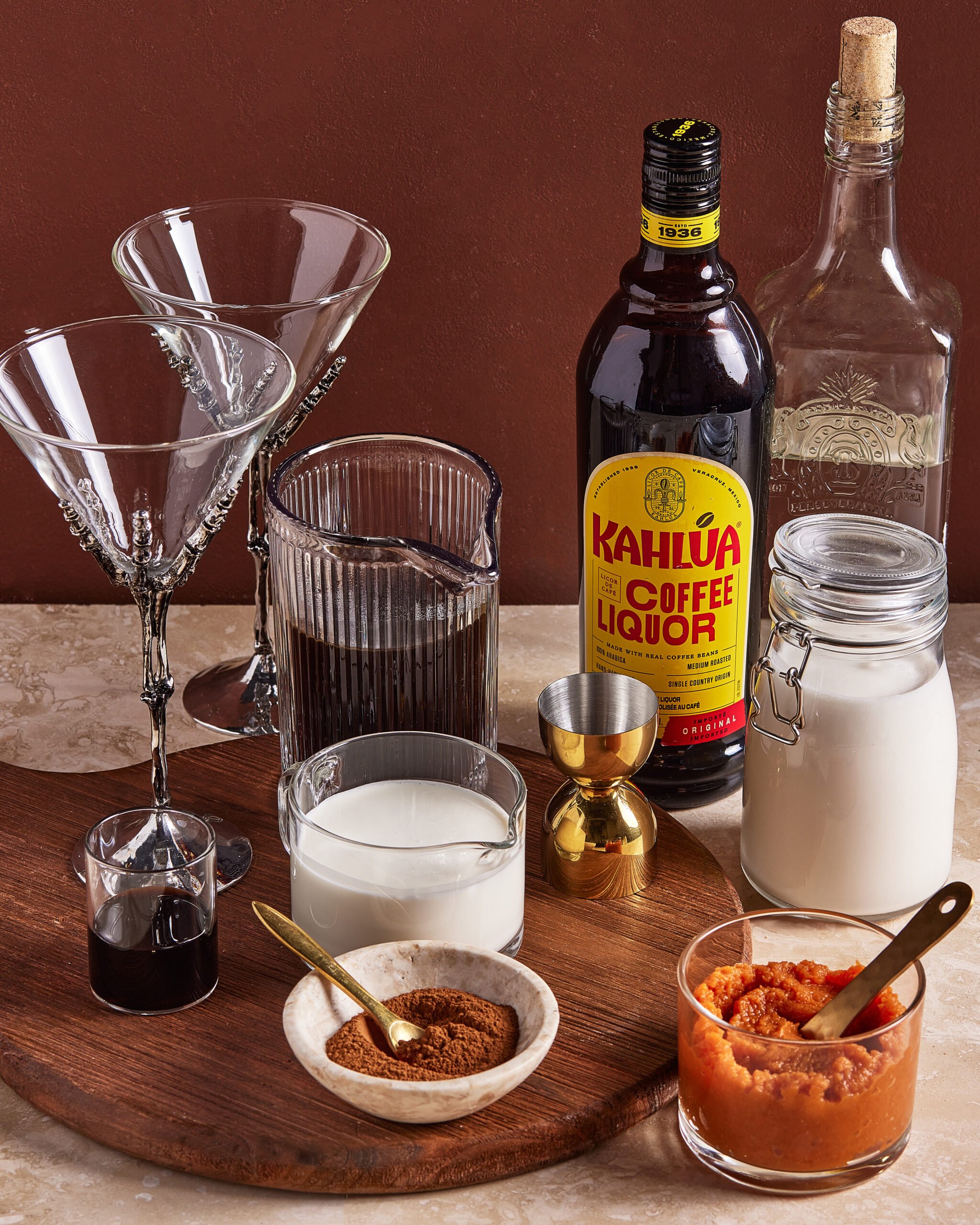A wooden board holds cocktail glasses, a bottle of Kahlúa, a glass bottle, milk, cream, a glass of dark liquid, a small gold jigger, a jar of puree, and a dish of cocoa powder against a brown background.