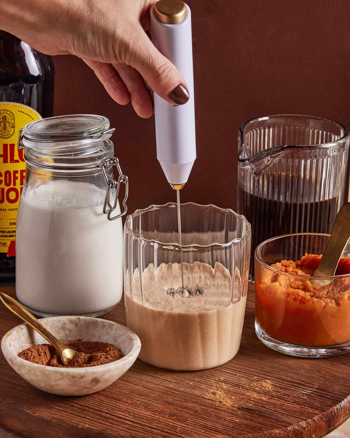 A hand uses a handheld frother to mix a creamy drink in a glass. Nearby are jars of milk, coffee, a bottle labeled "coffee liqueur," a small bowl of spice, and a bowl of orange puree on a wooden surface.