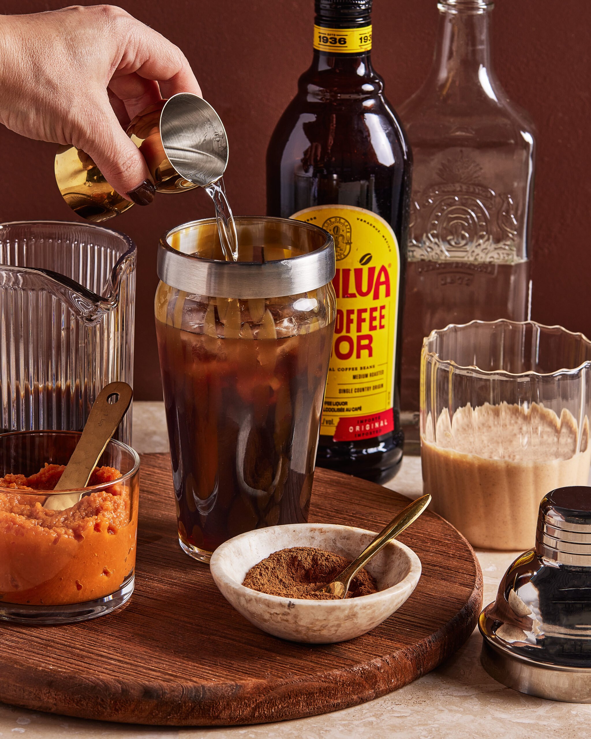 A hand pours liquid from a jigger into an iced coffee drink on a wooden tray, surrounded by Kahlúa bottle, glass pitcher, pumpkin puree, spices, cream, and a cocktail shaker.