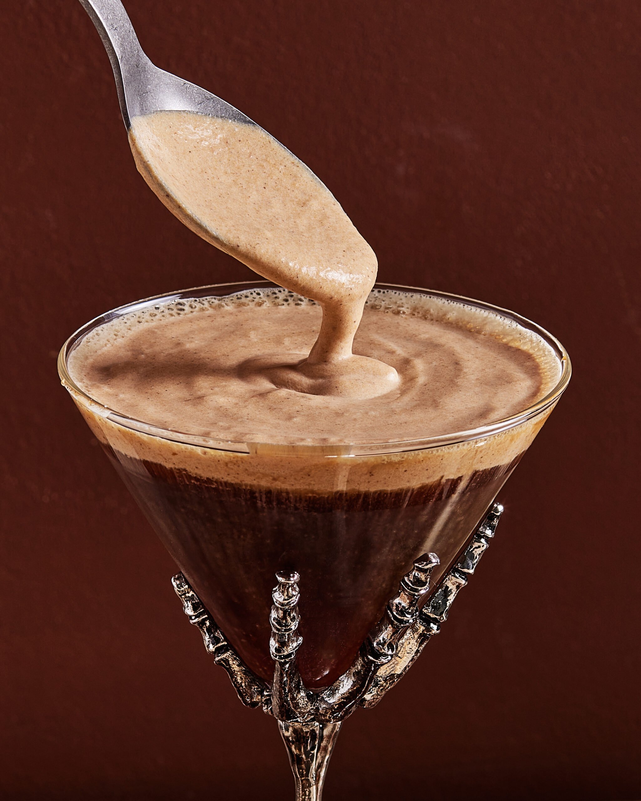 A close-up of a creamy brown liquid being poured from a spoon into a martini glass with a decorative skeletal hand stem, set against a dark brown background.