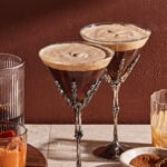 Two espresso martinis in ornate skeleton-hand glasses on a table, surrounded by glasses containing orange puree, brown liquid, and cinnamon powder, against a reddish-brown wall.