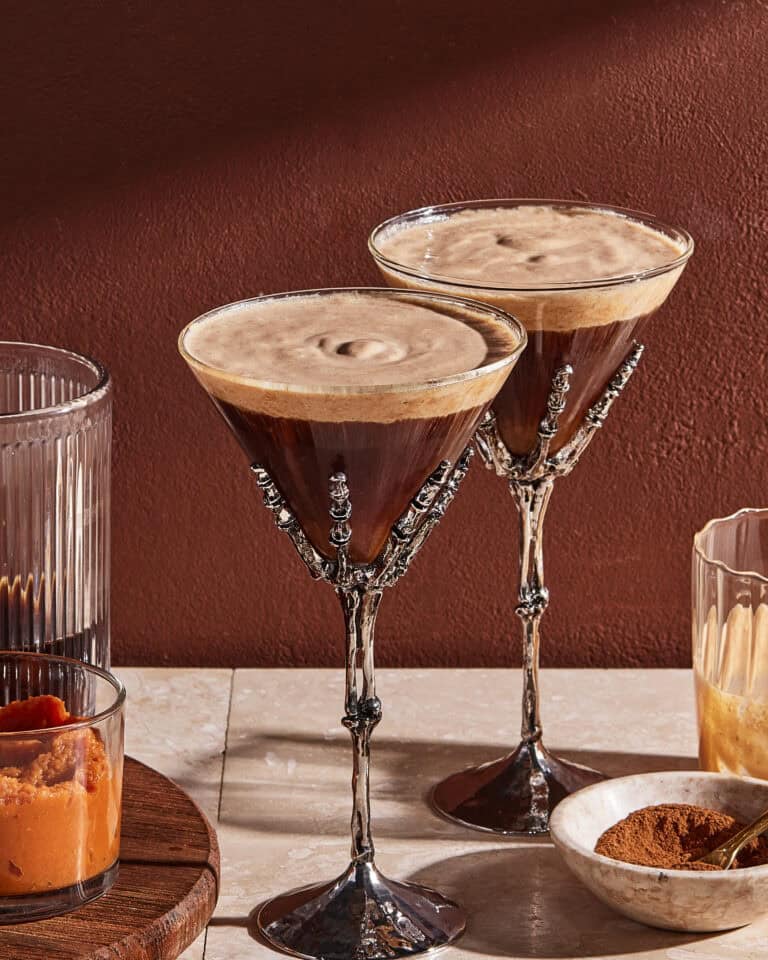 Two espresso martinis in ornate skeleton-hand glasses on a table, surrounded by glasses containing orange puree, brown liquid, and cinnamon powder, against a reddish-brown wall.