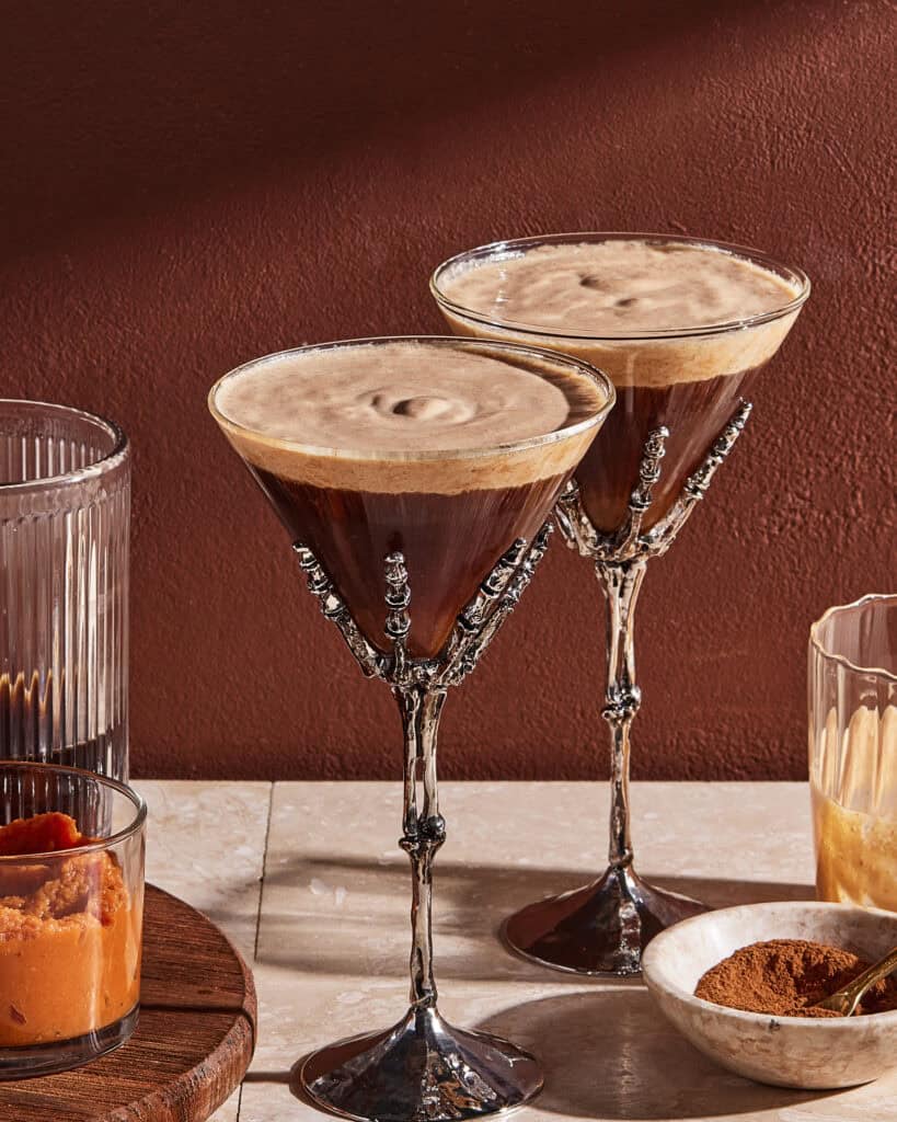 Two espresso martinis in ornate skeleton-hand glasses on a table, surrounded by glasses containing orange puree, brown liquid, and cinnamon powder, against a reddish-brown wall.