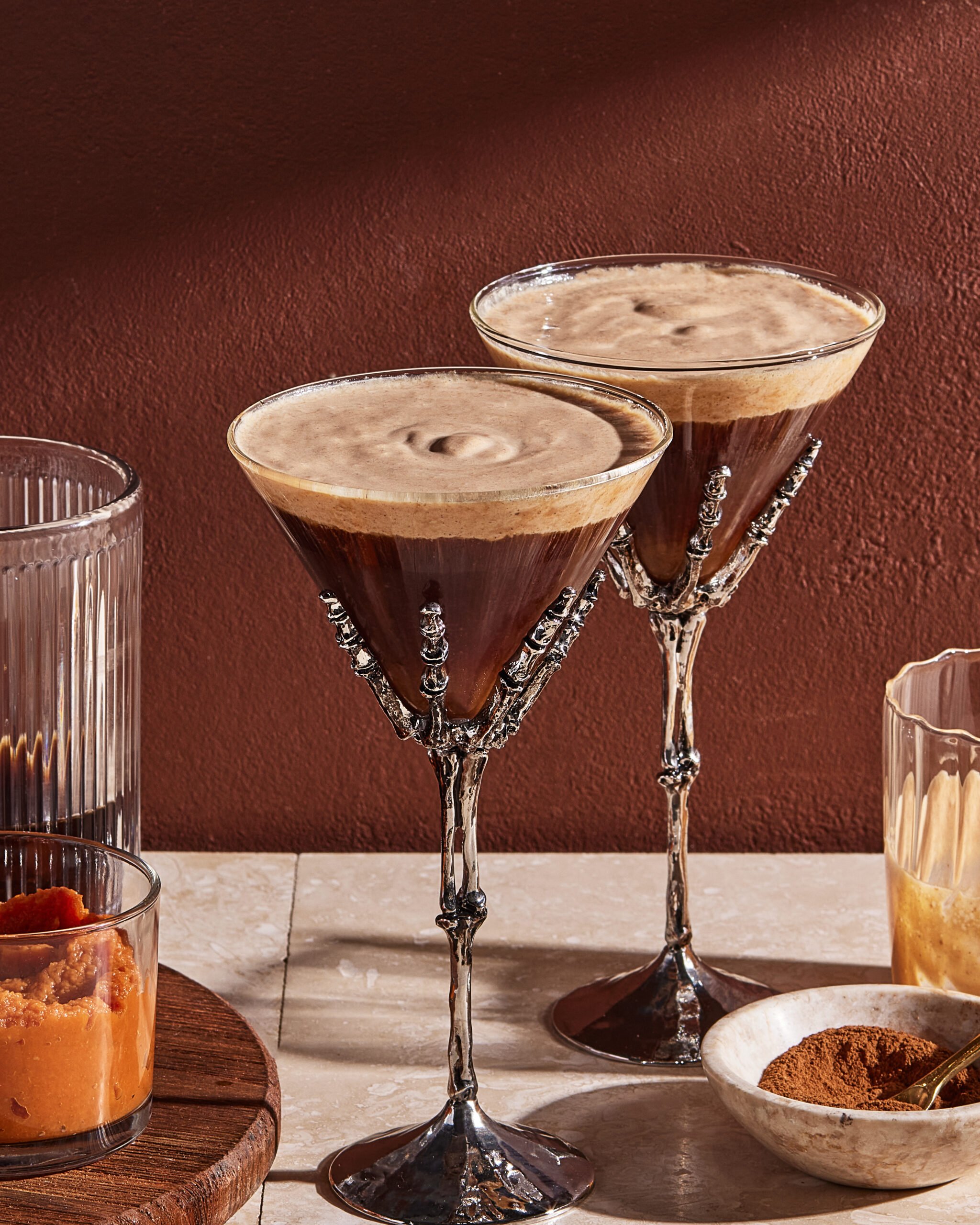 Two espresso martinis in ornate skeleton-hand glasses on a table, surrounded by glasses containing orange puree, brown liquid, and cinnamon powder, against a reddish-brown wall.