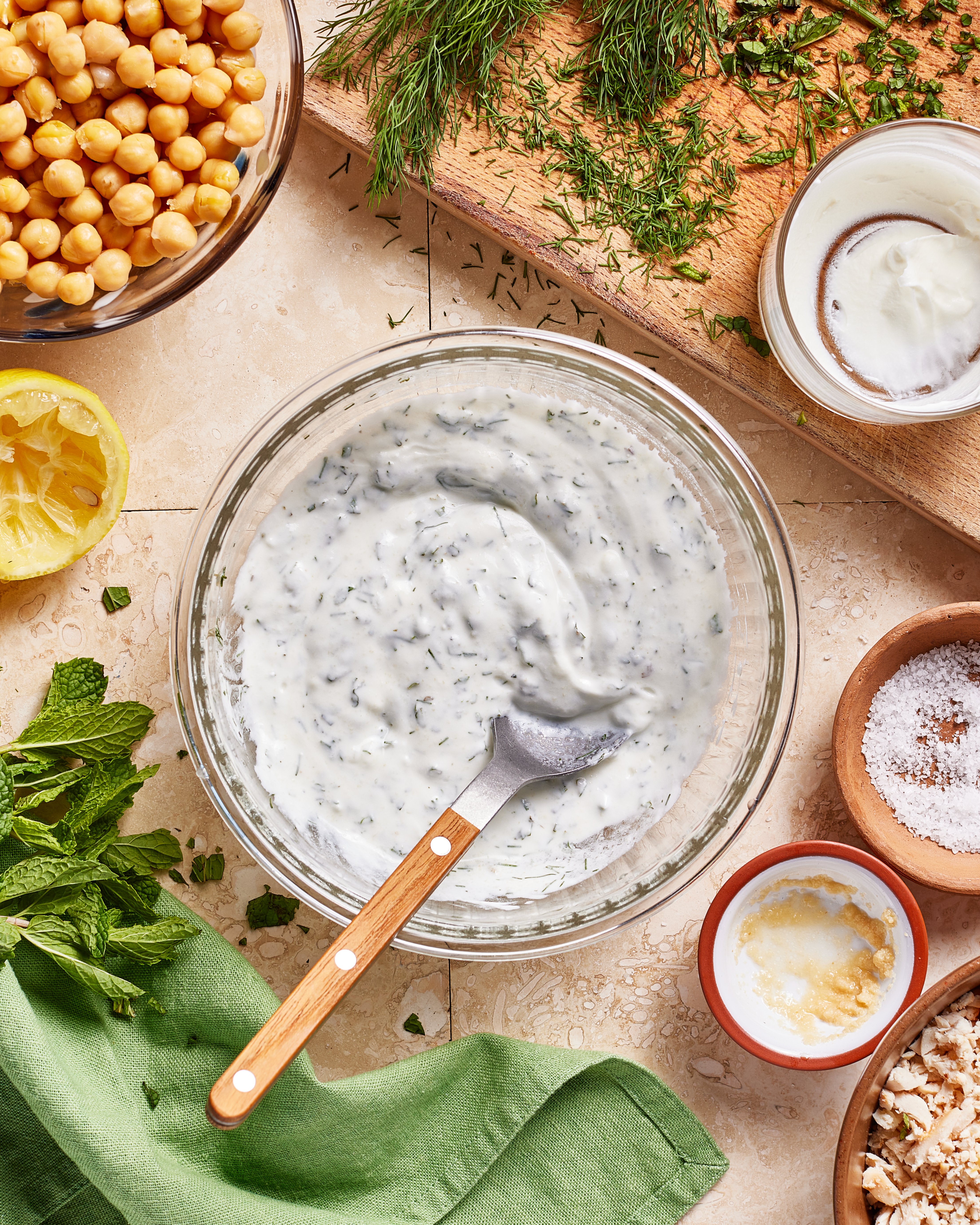 A bowl of herbed yogurt dip with a spoon sits on a tiled surface, surrounded by fresh herbs, chickpeas, a lemon half, grated garlic, salt, and a green napkin.