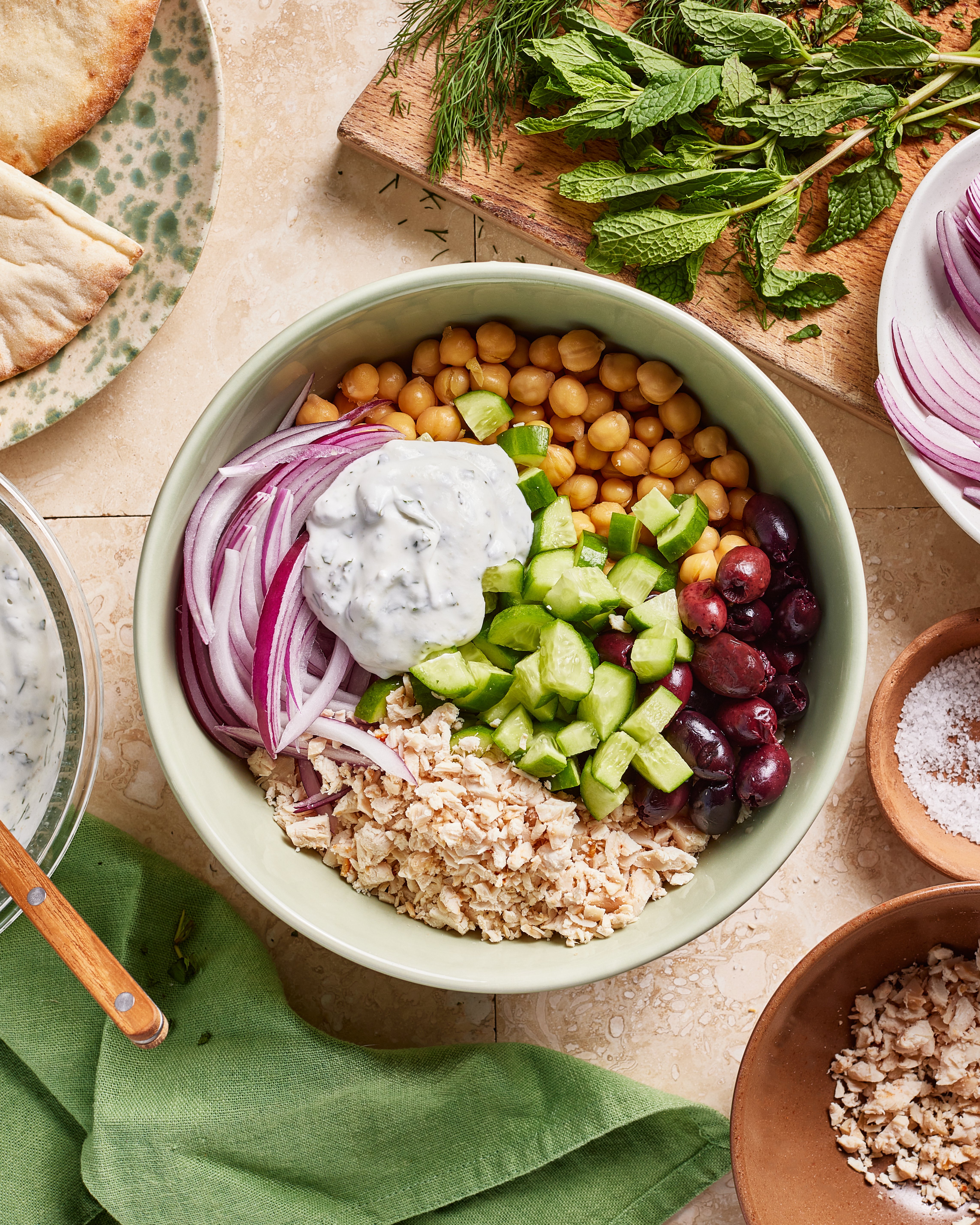 A bowl filled with chopped cucumber, red onion, chickpeas, kalamata olives, crumbled pita, and tzatziki sauce, surrounded by fresh herbs, pita bread, and other ingredients on a table.