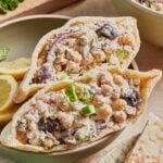Two pita halves filled with a chickpea salad containing cucumber, red onion, black olives, herbs, and a creamy dressing, served on a plate with lemon slices and pita bread on the side.