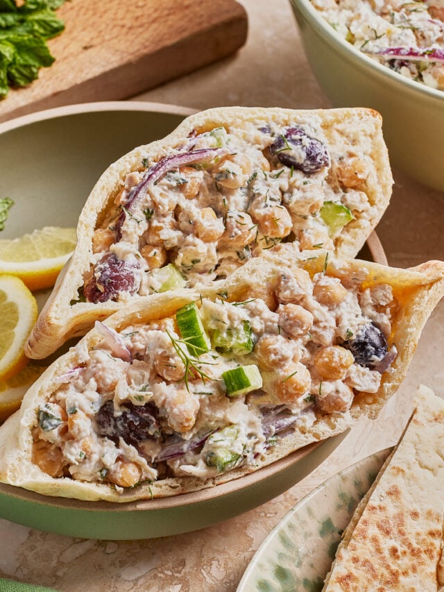 Two pita halves filled with a chickpea salad containing cucumber, red onion, black olives, herbs, and a creamy dressing, served on a plate with lemon slices and pita bread on the side.
