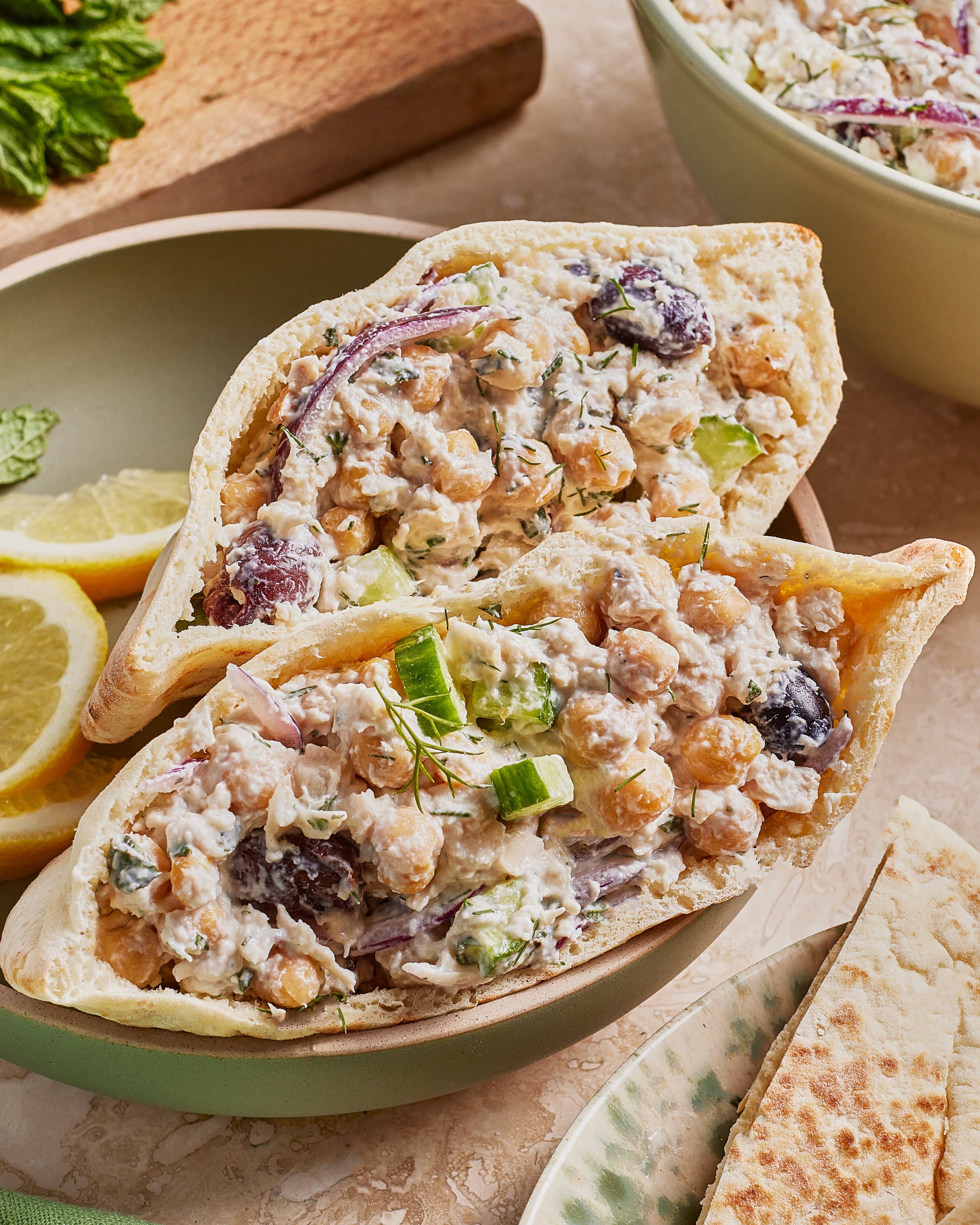 Two pita halves filled with a chickpea salad containing cucumber, red onion, black olives, herbs, and a creamy dressing, served on a plate with lemon slices and pita bread on the side.