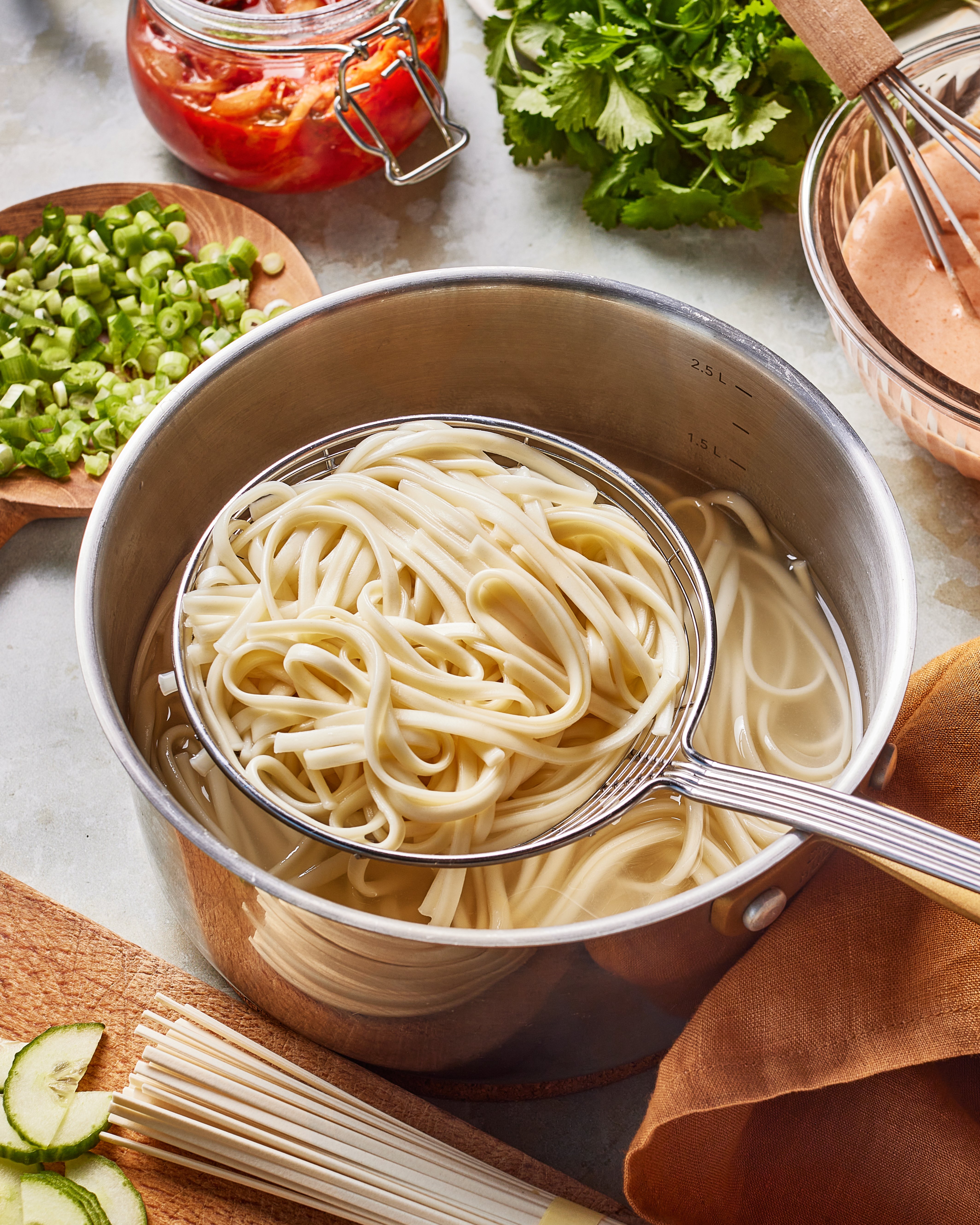 A metal pot filled with cooked noodles in water sits on a counter. A slotted spoon lifts some noodles out. Chopped green onions, cilantro, a jar of pickled vegetables, and sauce are nearby.