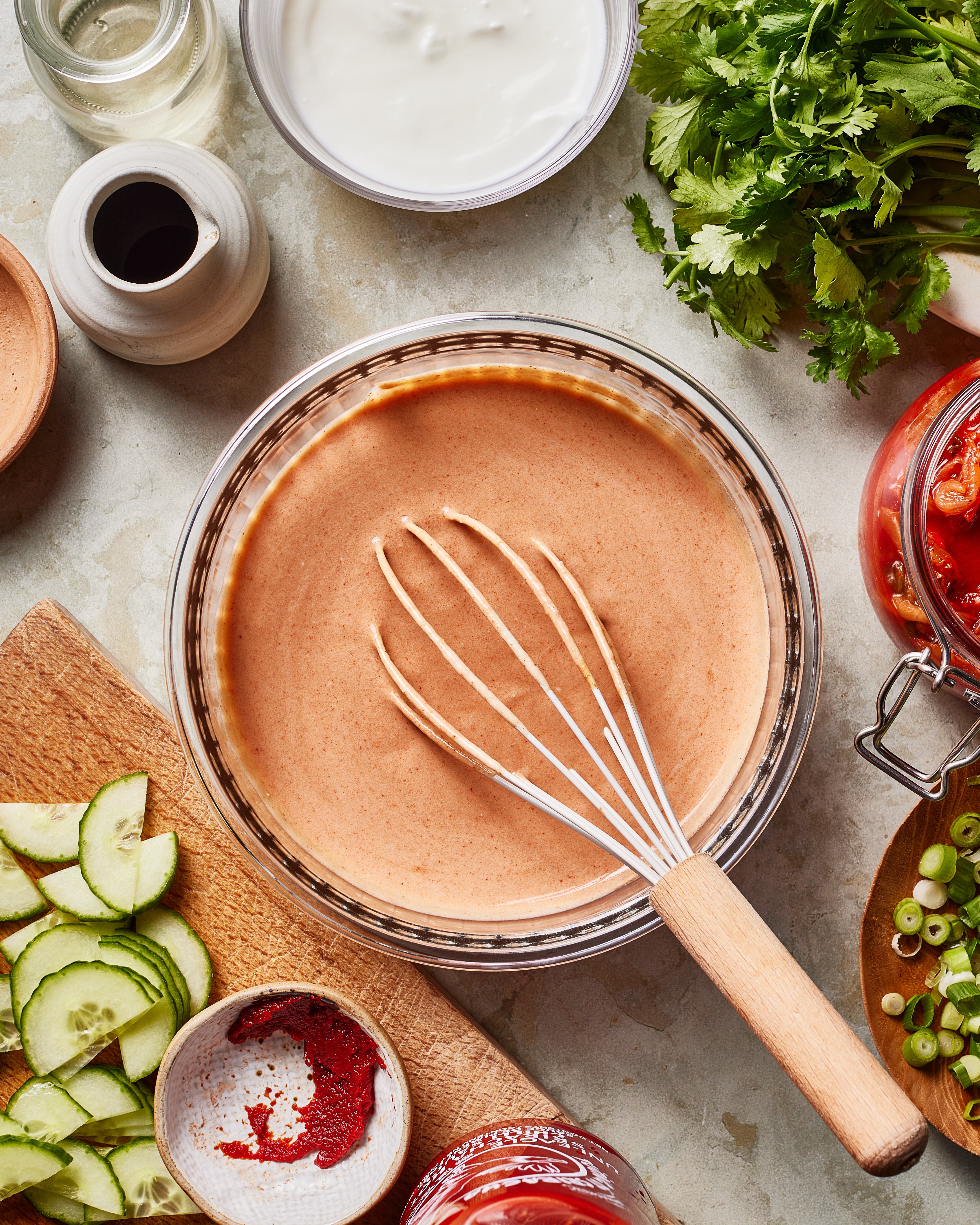 A glass bowl filled with creamy pink sauce and a whisk sits on a countertop surrounded by sliced cucumbers, fresh herbs, chopped green onions, a small bowl of red spice, and jars and bowls with various ingredients.