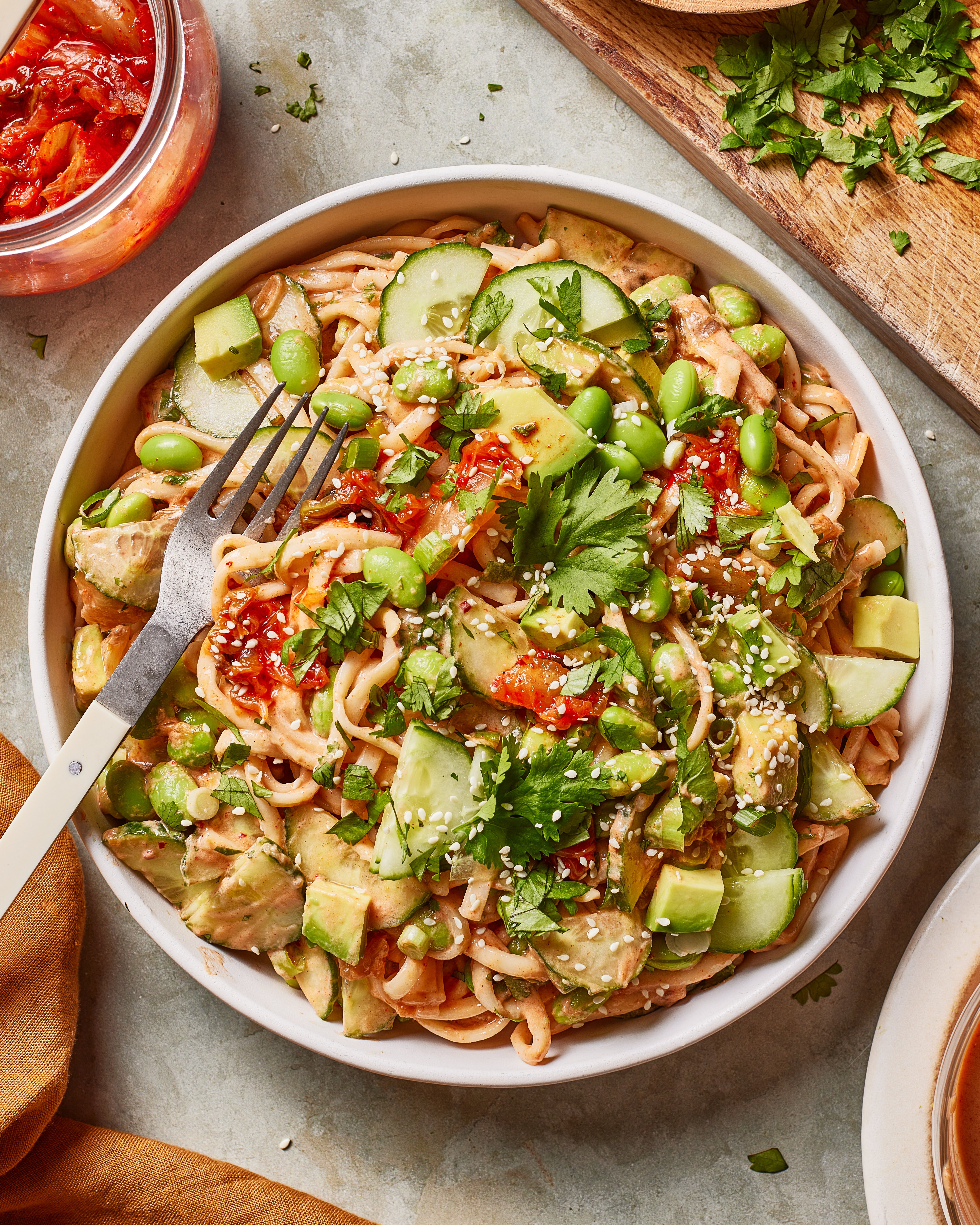 A bowl of noodles topped with cubed avocado, cucumber slices, edamame, sun-dried tomatoes, cilantro, and sesame seeds, with a fork resting on the bowl. Fresh herbs and condiments are visible in the background.