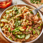 These udon noodle bowls are topped with avocado slices, cucumber, edamame, cilantro, and sesame seeds. A fork is twirling noodles, and a jar of chili sauce and a cutting board with chopped herbs are nearby.