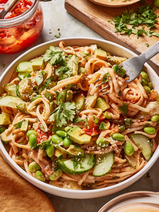 These udon noodle bowls are topped with avocado slices, cucumber, edamame, cilantro, and sesame seeds. A fork is twirling noodles, and a jar of chili sauce and a cutting board with chopped herbs are nearby.