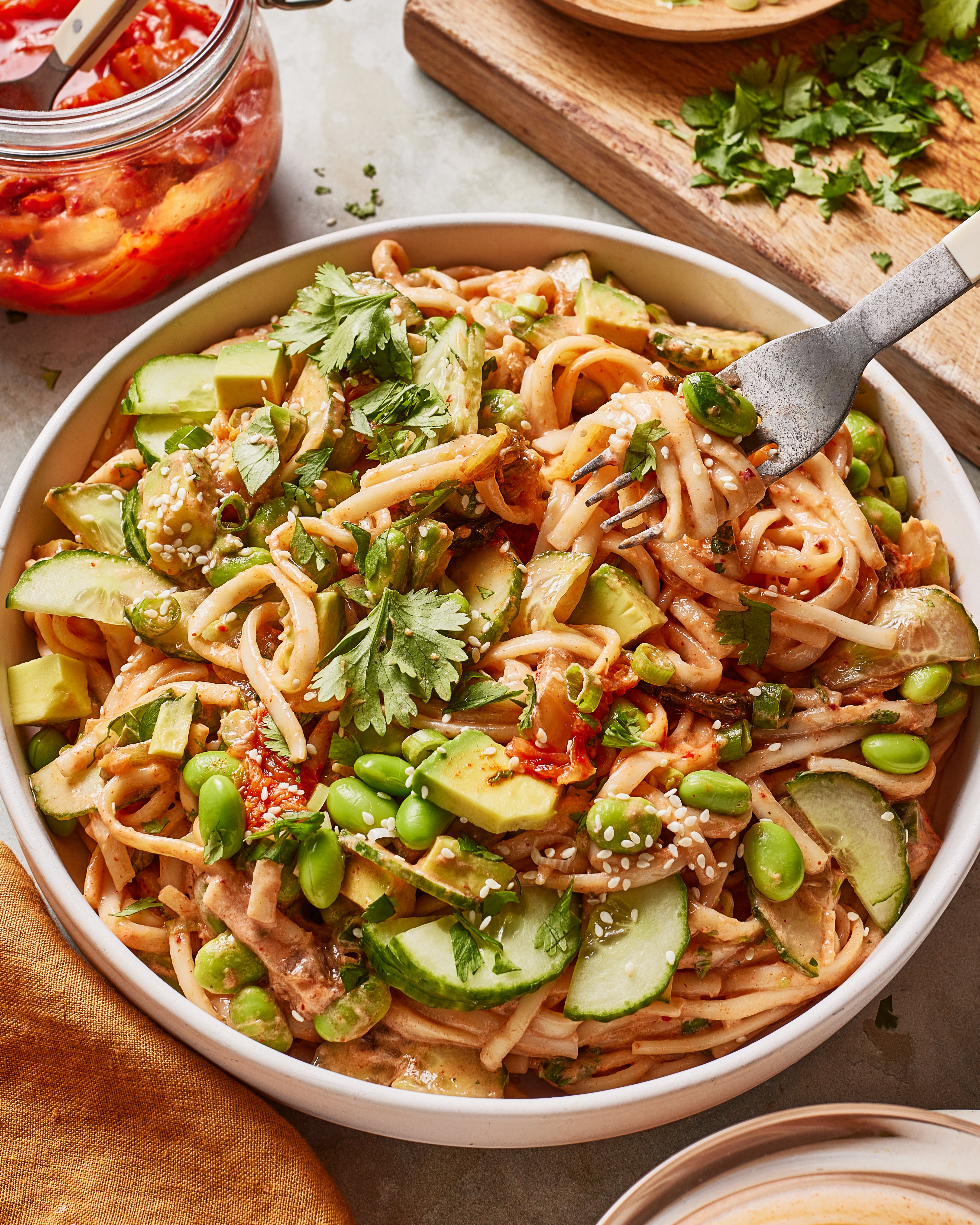 These udon noodle bowls are topped with avocado slices, cucumber, edamame, cilantro, and sesame seeds. A fork is twirling noodles, and a jar of chili sauce and a cutting board with chopped herbs are nearby.