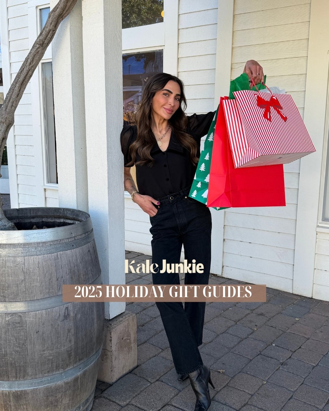 A woman in black clothing stands outside a white building, smiling and holding colorful holiday gift bags. Text on the image reads “Kale Junkie 2025 Holiday Gift Guides.”.