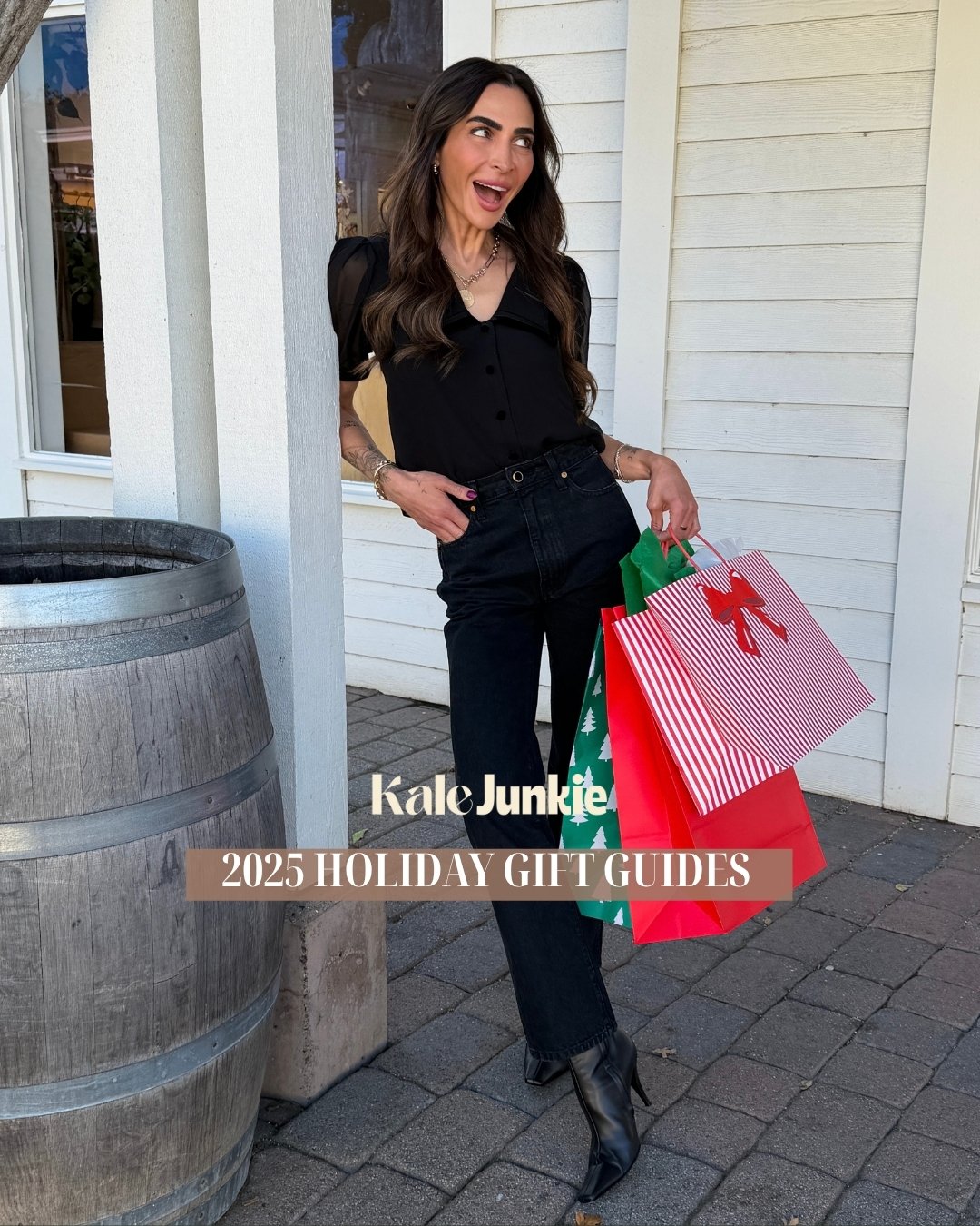 A woman with long dark hair, wearing a black blouse and jeans, stands smiling outside near a barrel, holding holiday gift bags. Overlay text reads “Kale Junkie” and “2025 Holiday Gift Guides.”.