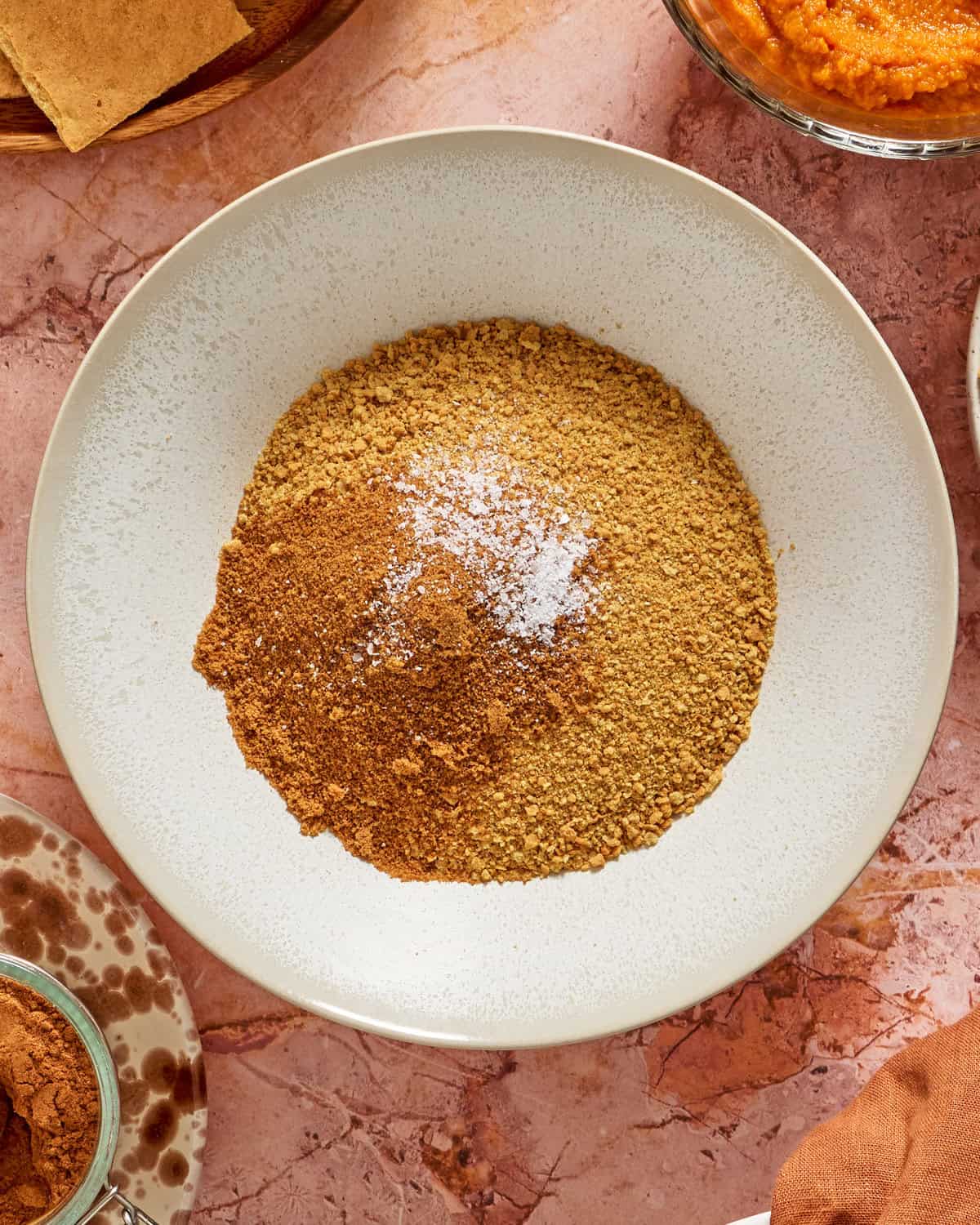 A white bowl filled with ground graham crackers, cinnamon, and a pinch of salt sits on a marble countertop, surrounded by bowls with pumpkin puree, cinnamon, and other ingredients.