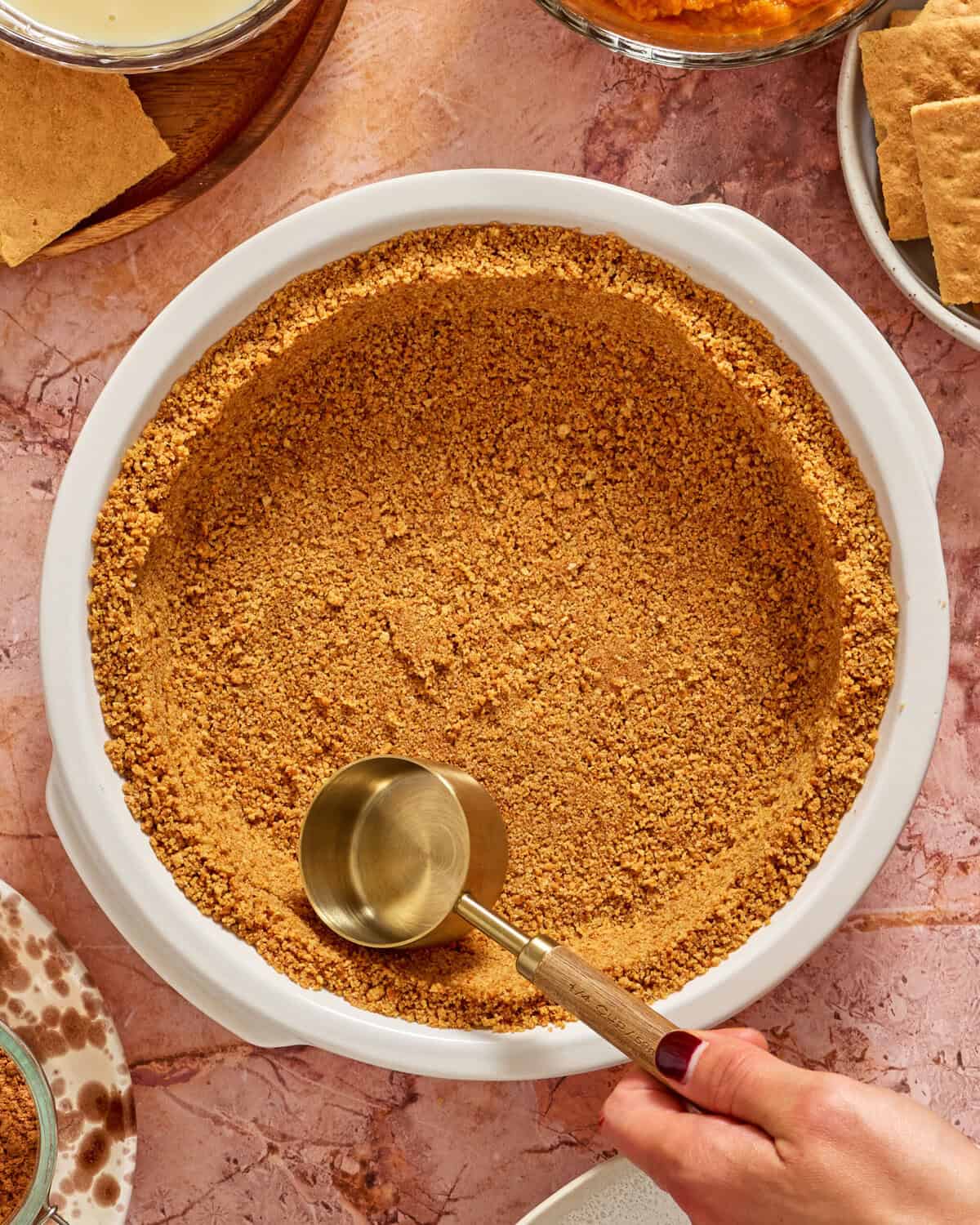 A hand presses graham cracker crumbs into a white pie dish using a metal measuring cup, surrounded by graham crackers and dessert ingredients on a pink marble surface.