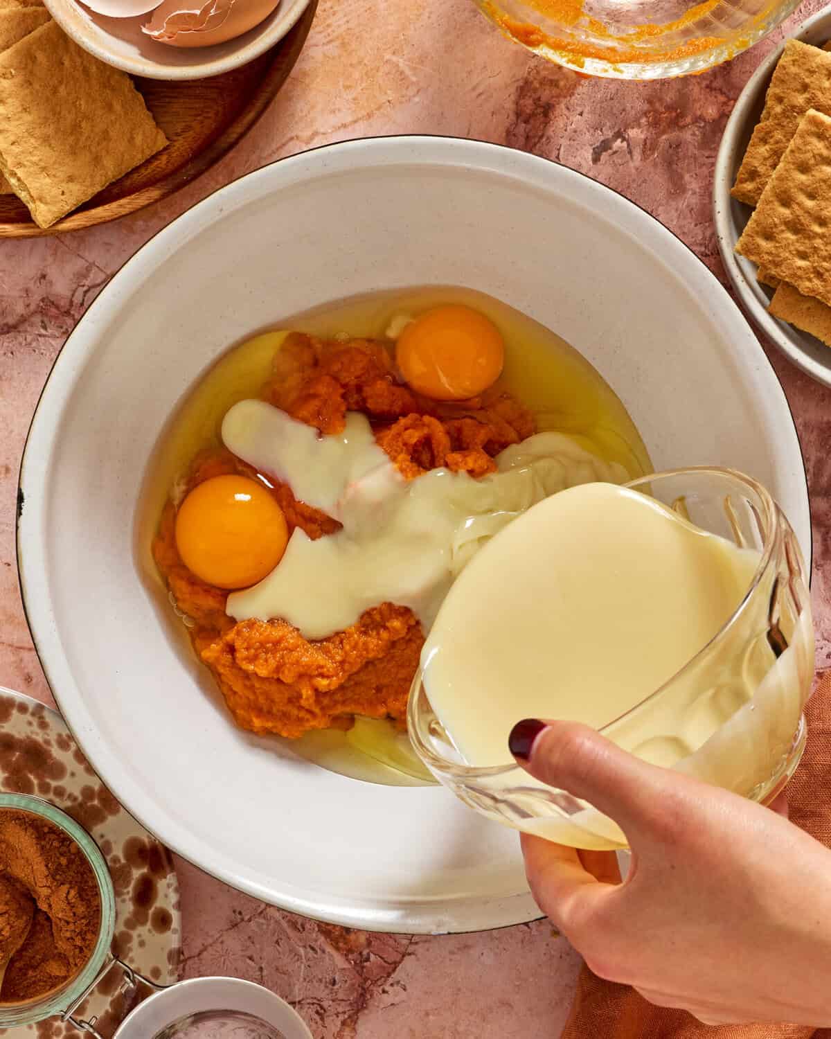 A hand pours sweetened condensed milk into a large bowl containing eggs and pumpkin puree, surrounded by graham crackers, cinnamon, and an empty eggshell on a pink marble surface.