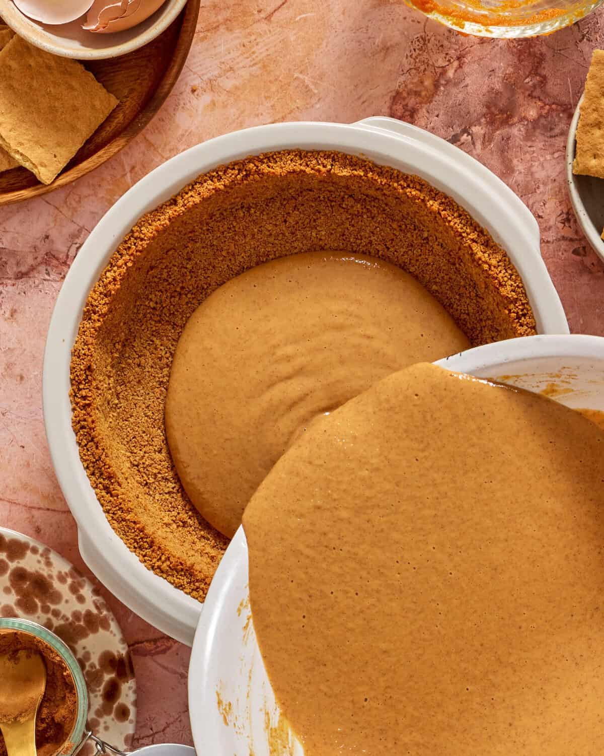 A creamy brown filling is being poured into a graham cracker crust in a white pie dish, surrounded by baking ingredients and utensils on a pink marble countertop.
