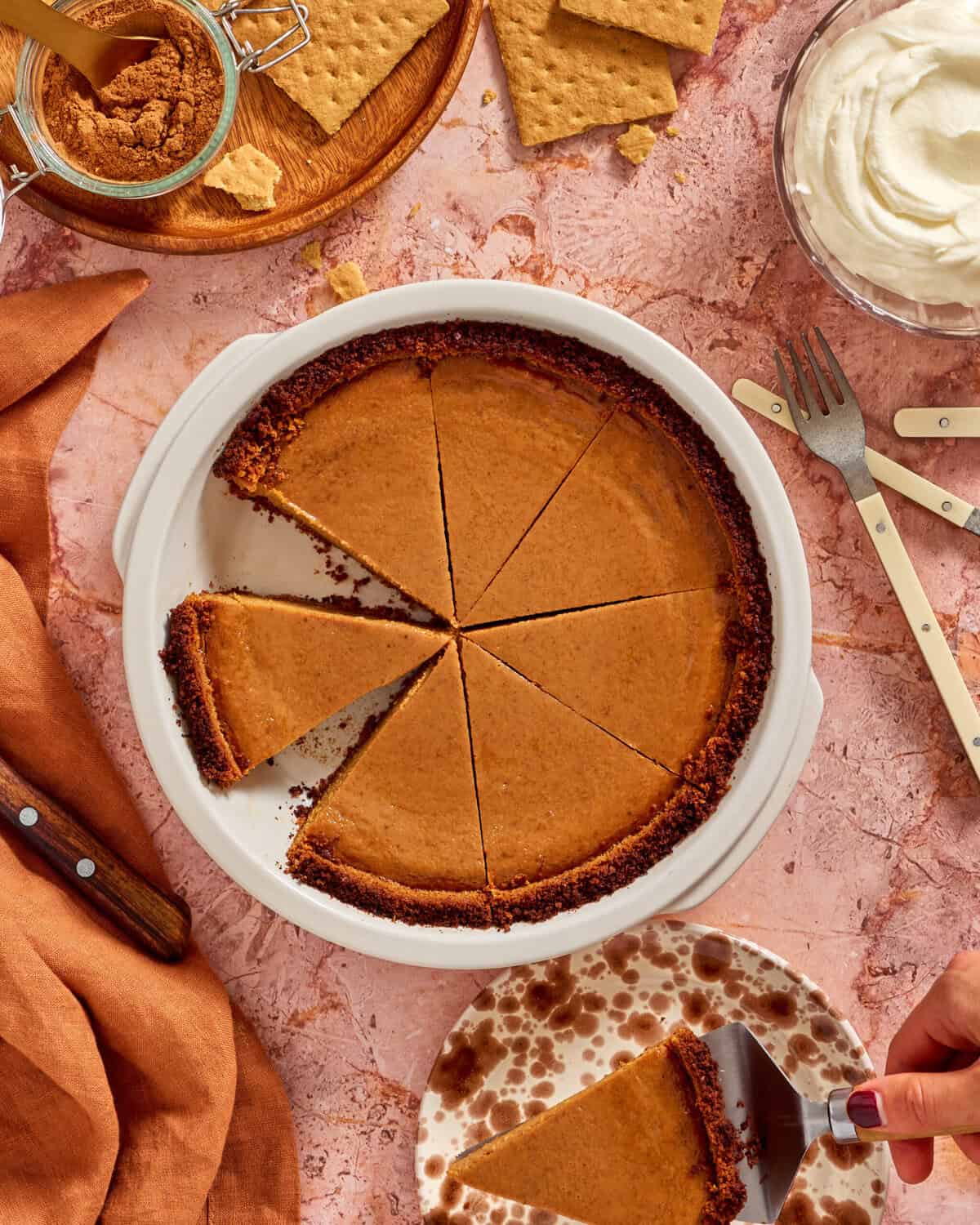 Top-down view of a sliced pumpkin pie in a white dish, with one slice being served onto a speckled plate. Surrounding items include graham crackers, whipped cream, and pie utensils on a pink surface.