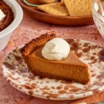 A slice of pumpkin pie with a graham cracker crust and a dollop of whipped cream sits on a decorative plate. Graham crackers, more pie, and a bowl of whipped cream are in the background.