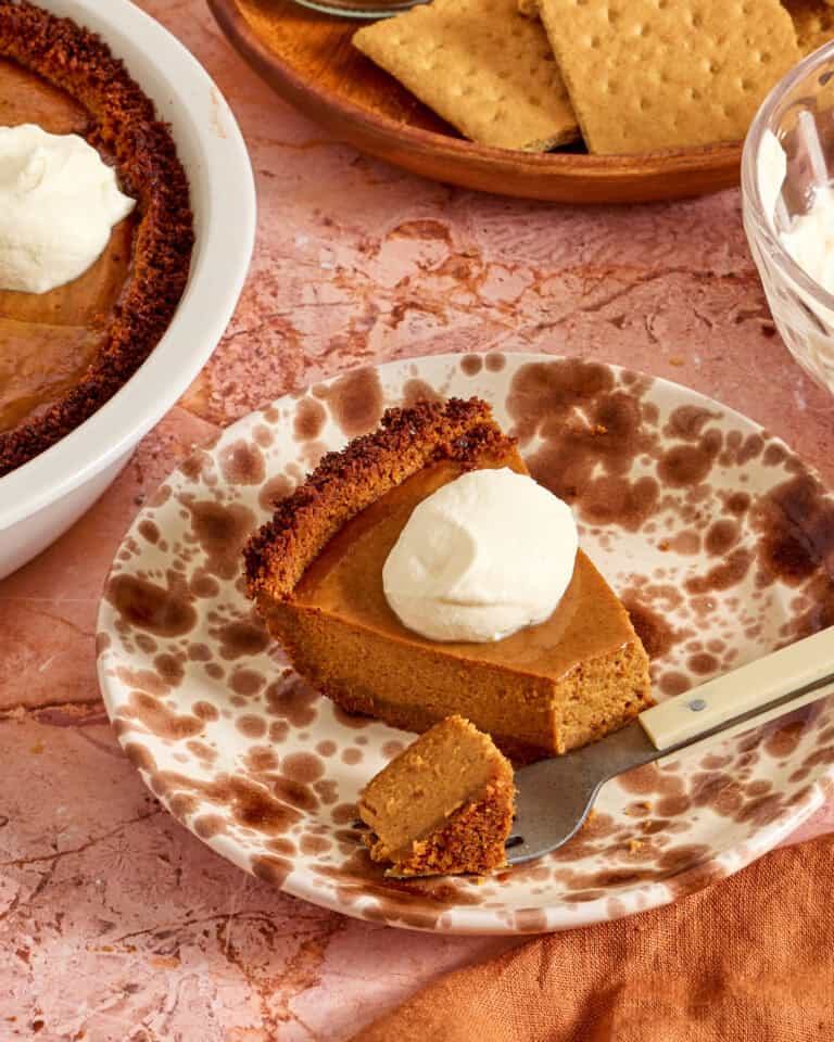 A slice of pumpkin pie with a graham cracker crust topped with whipped cream sits on a plate. A fork holds a small bite. In the background, the rest of the pie, more graham crackers, and a bowl of whipped cream are visible.
